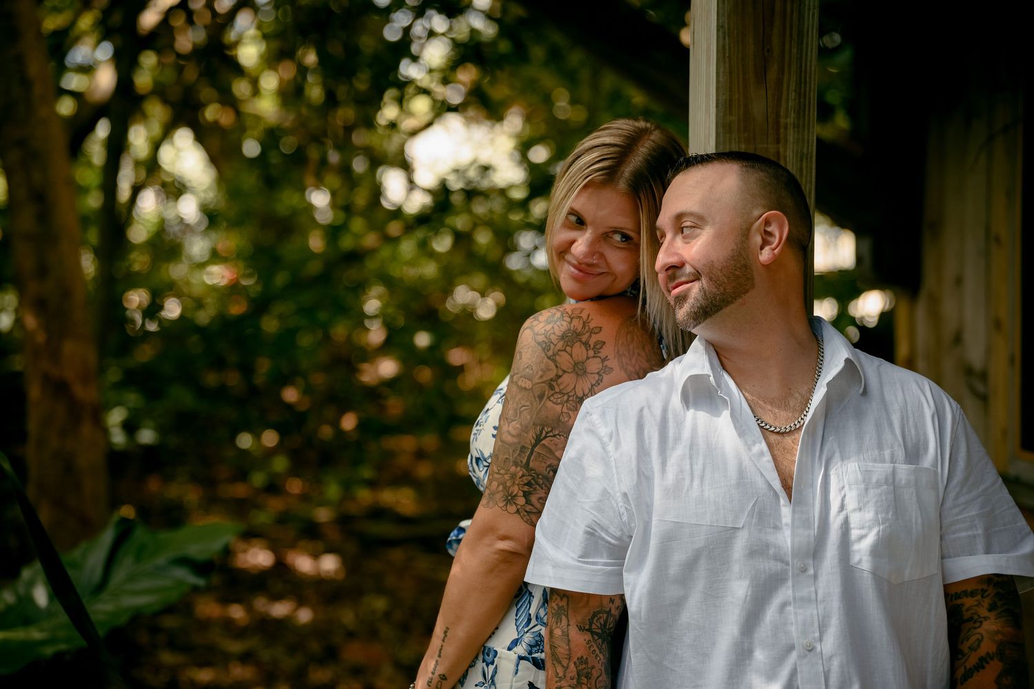 Tattooed couple posing outdoors in lush green setting, woman smiling over shoulder, man in white shirt looking away.