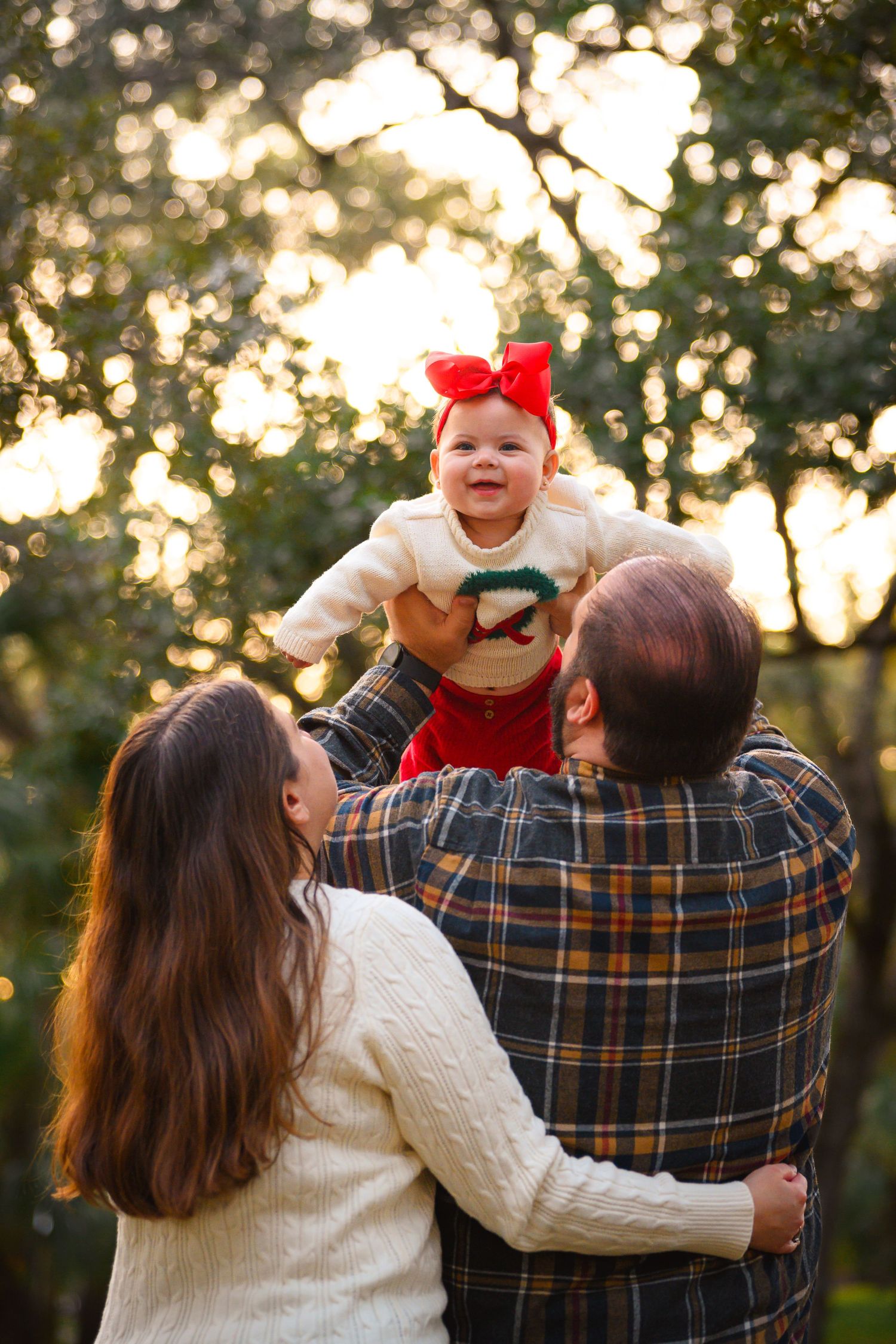 Parents lift smiling baby girl with red bow above their heads in golden park.