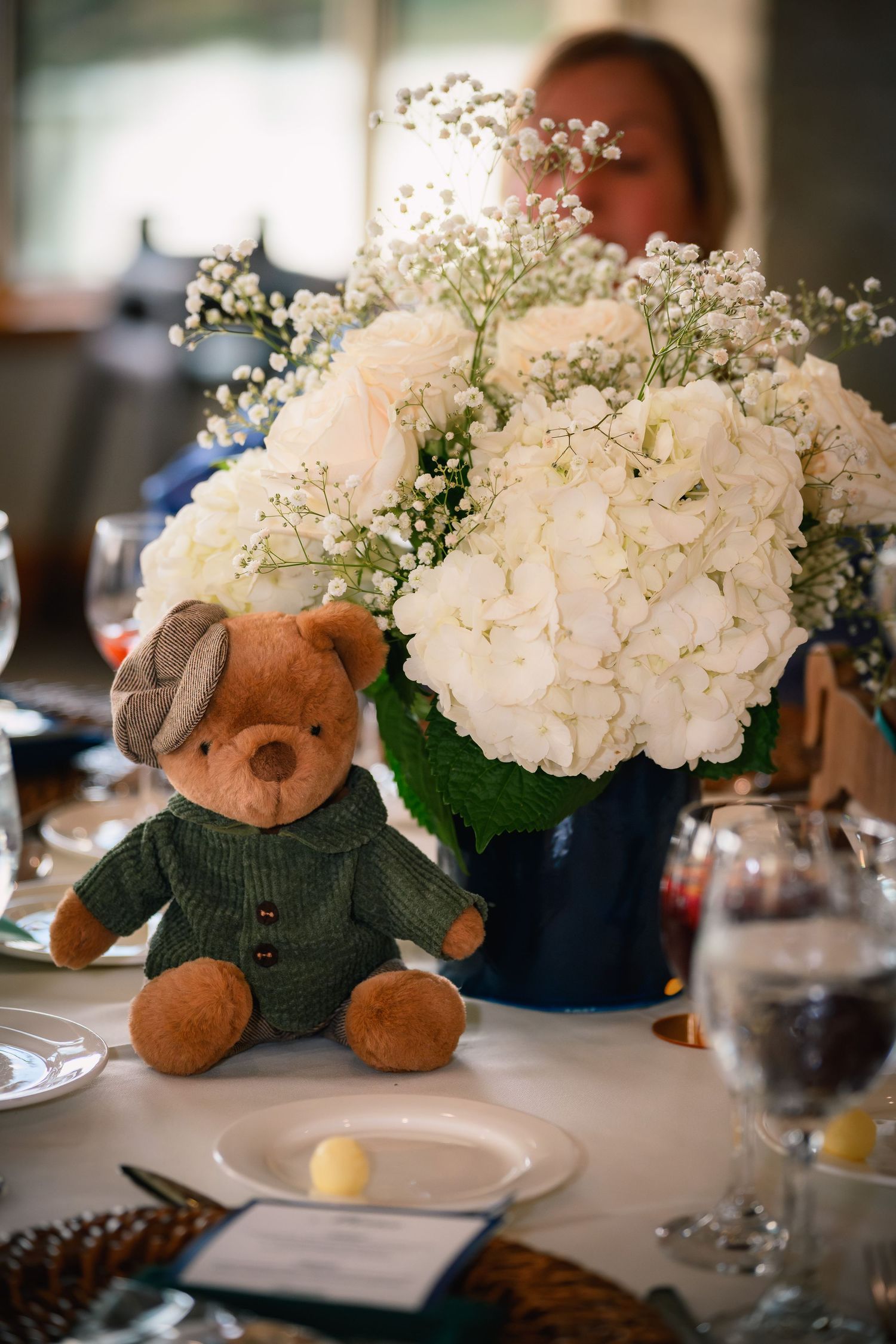 Adorable teddy bear in green sweater sits beside white hydrangea and rose floral centerpiece on decorated table.