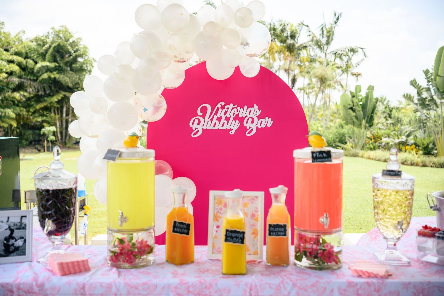 Colorful beverage station with glass drink dispensers and pink decorative backdrop at an outdoor party setup.