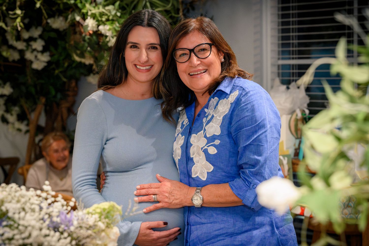 Pregnant woman and older woman smiling together at floral baby shower, hand on baby bump.