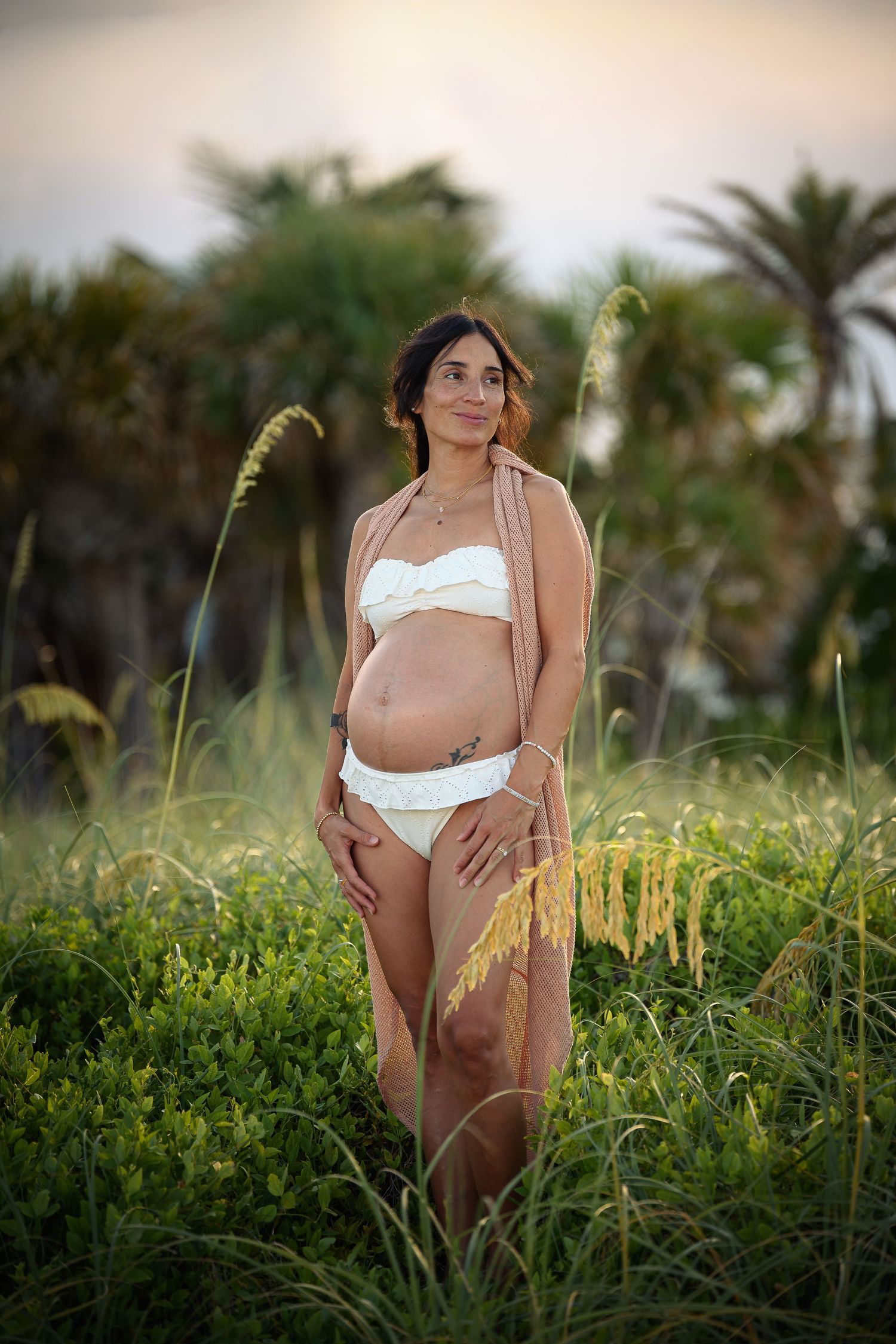 Pregnant woman in white bikini and shawl standing in lush green coastal vegetation with palm trees at sunset.