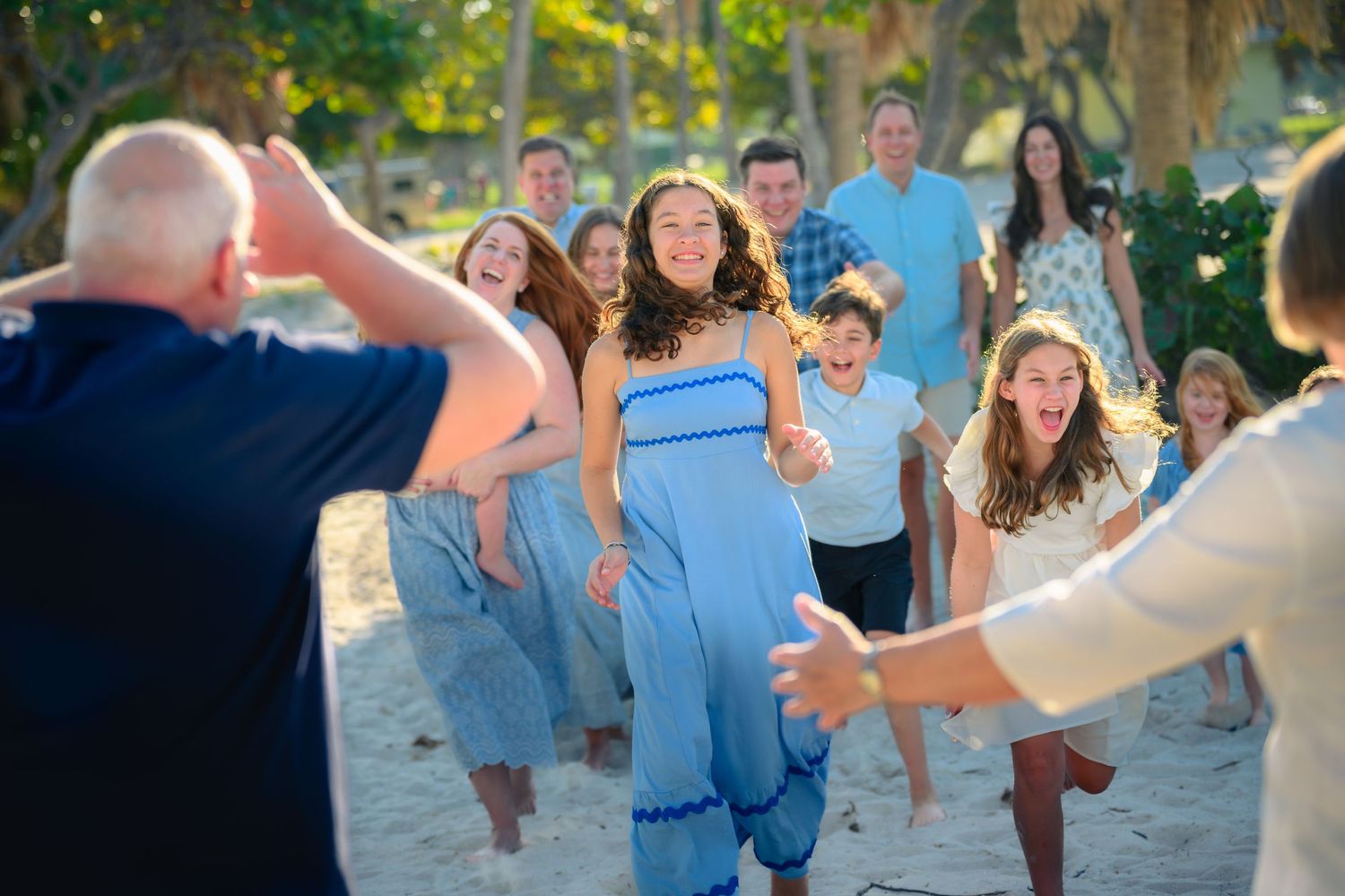 Family reunion on a sunny beach with a girl in a blue dress running toward open arms while others smile and cheer.