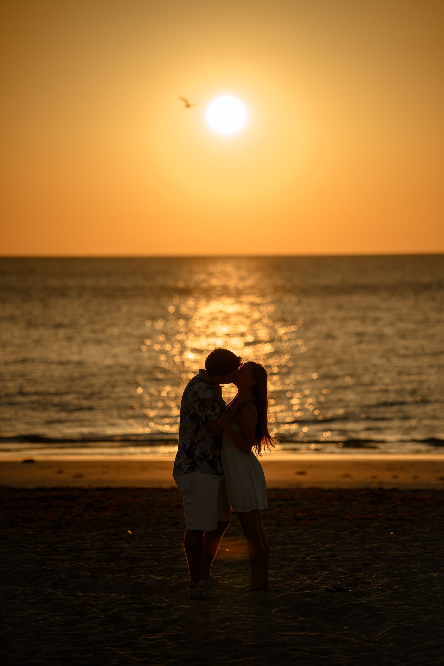 Romantic couple silhouette kissing on beach at golden sunset with ocean backdrop and glowing sun.