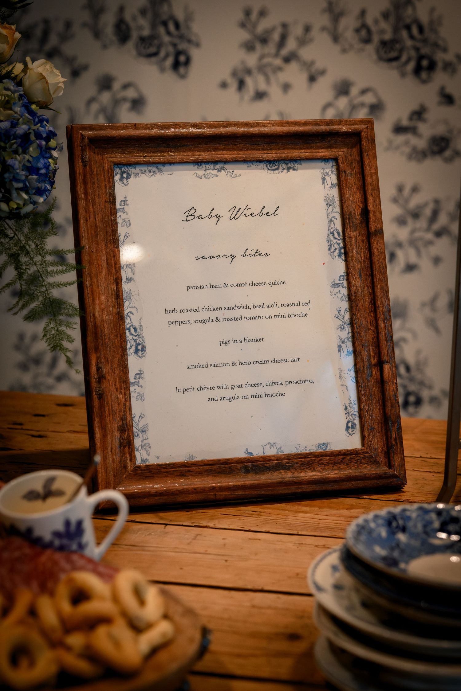 Rustic wooden frames display elegant text on tables with delicate blue and white floral wallpaper in the background.