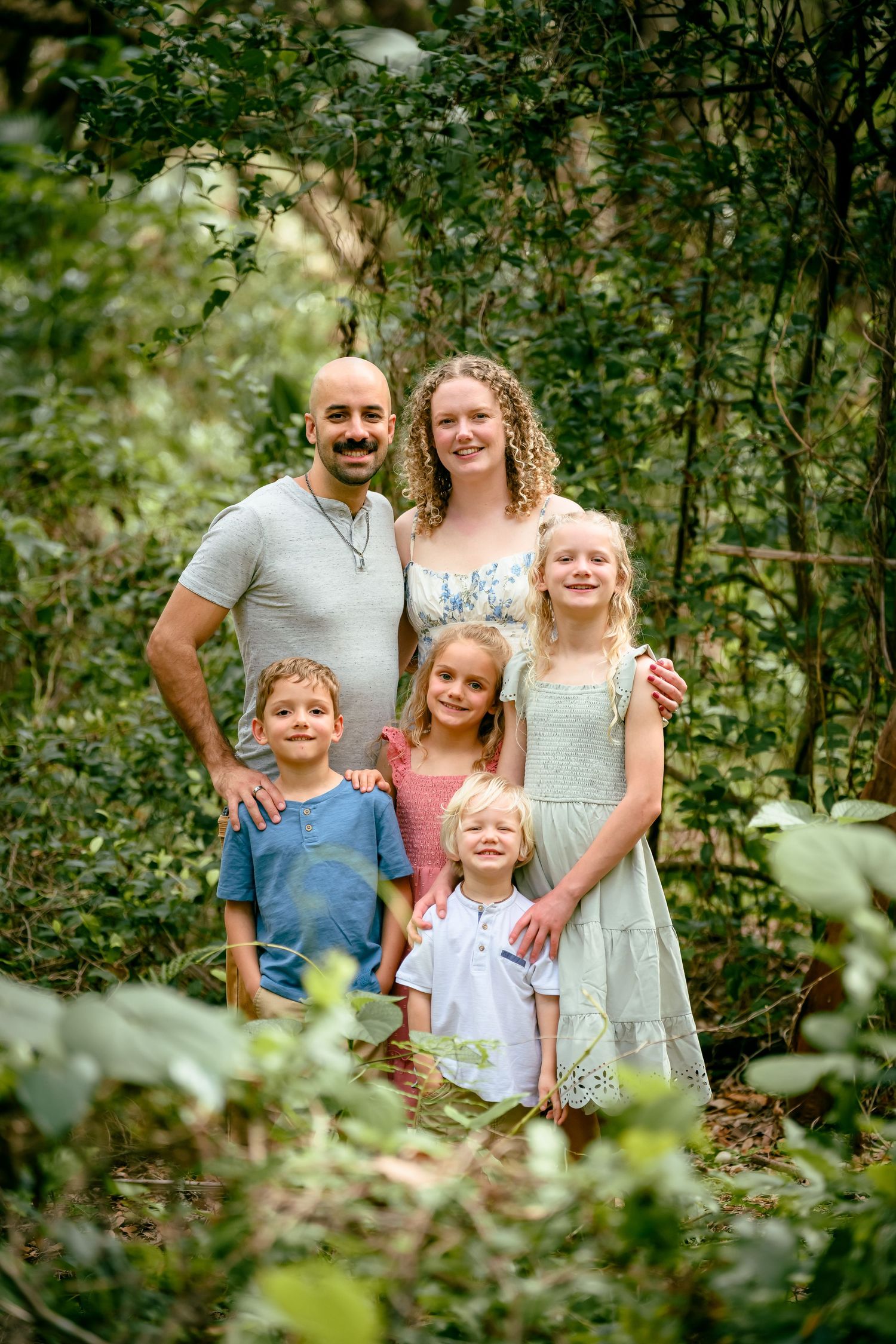 Family of six posing outdoors among lush green foliage, parents with four young children smiling together.