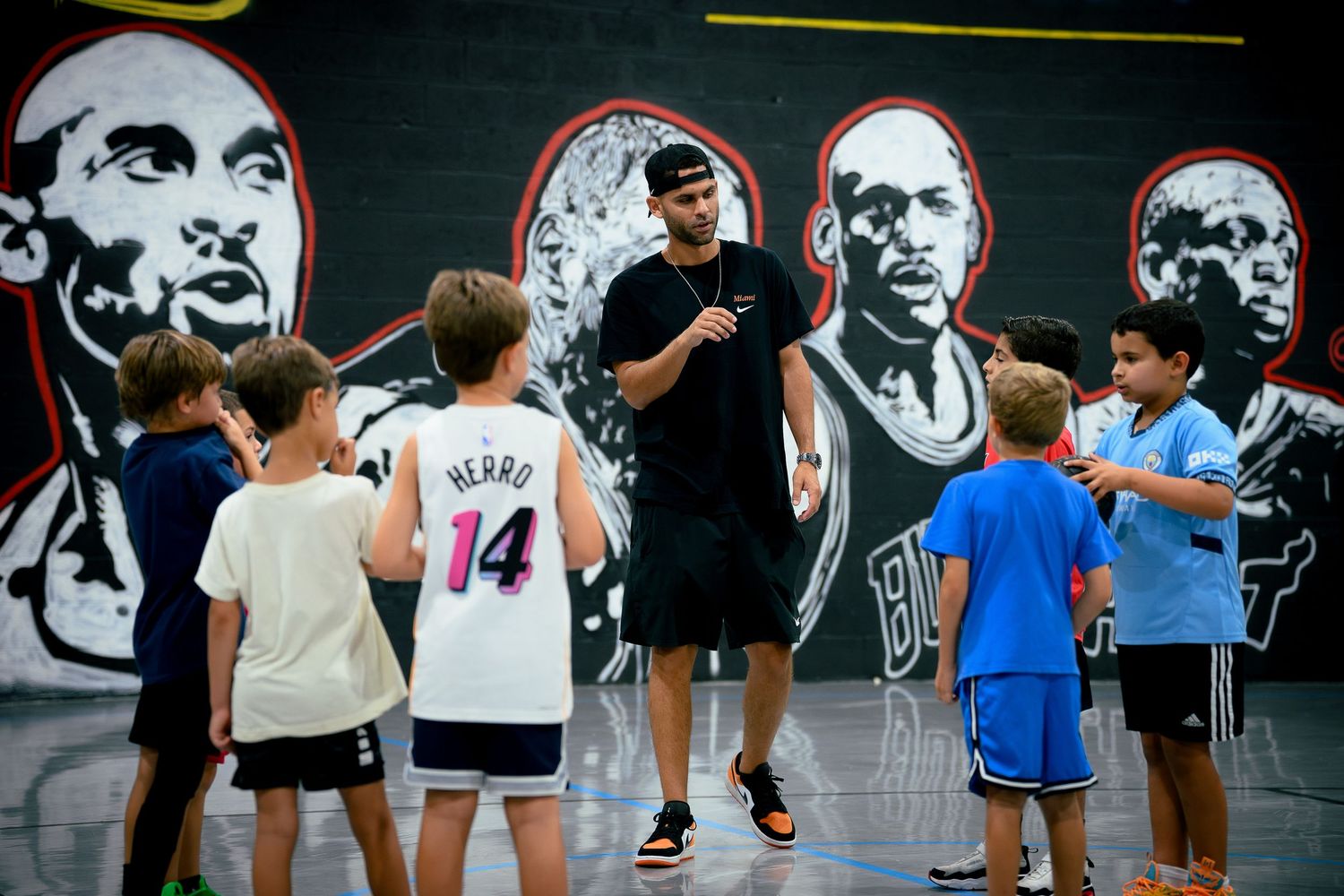 Basketball coach instructs group of young kids on indoor court with mural backdrop.