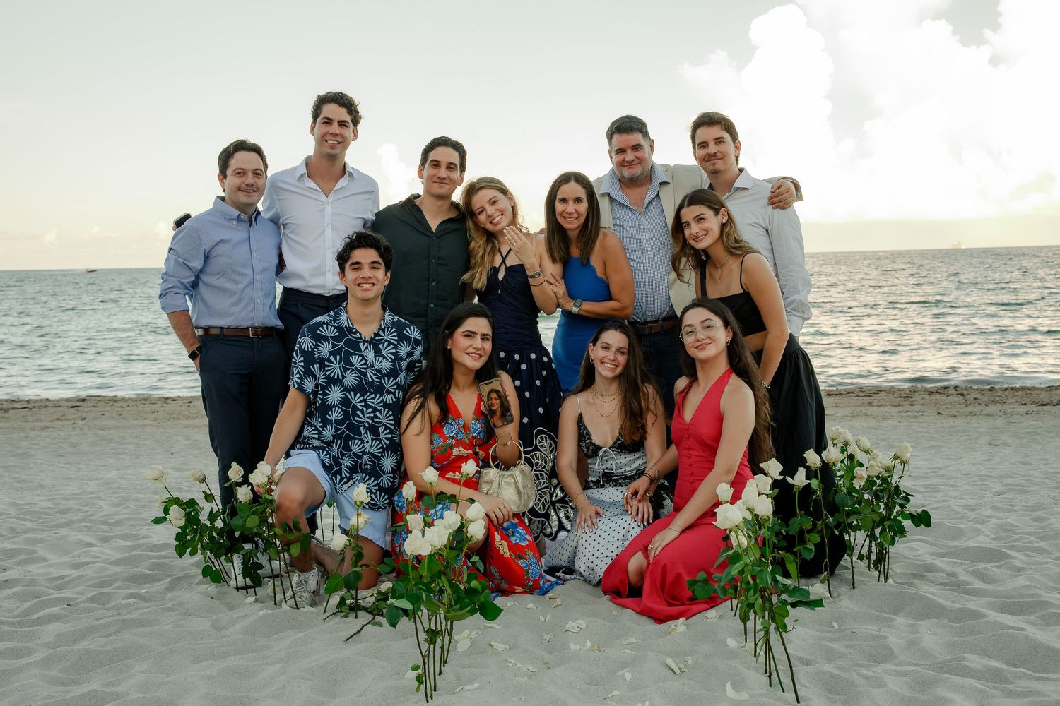 Group of young adults posing together on a beach at sunset with white roses arranged in the foreground.