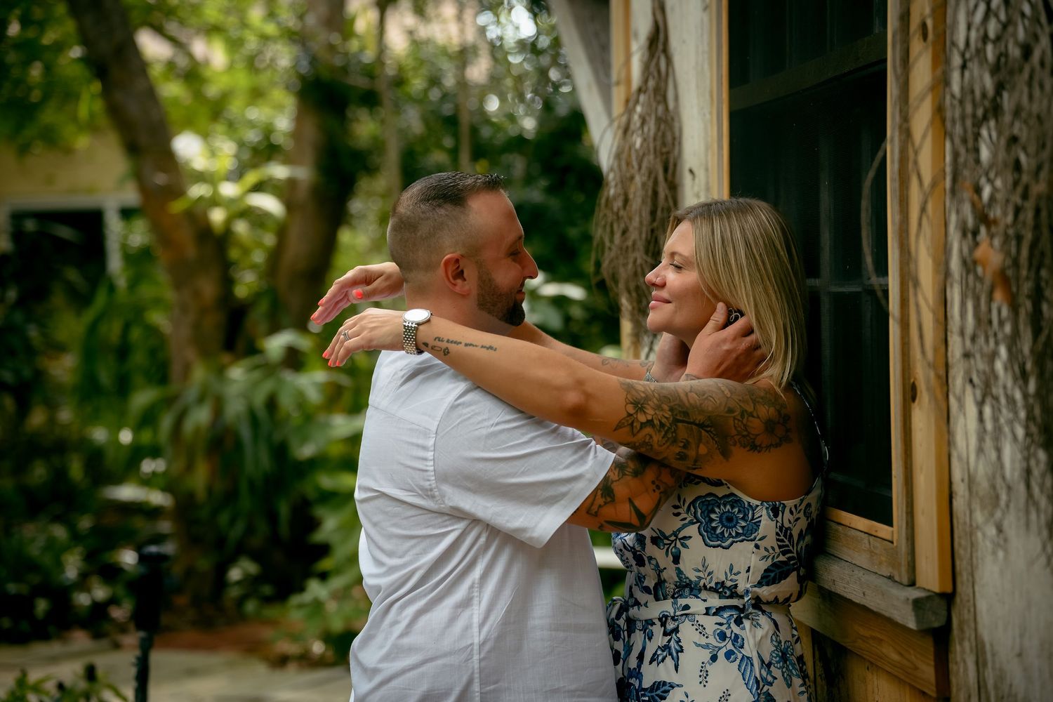 Couple sharing a romantic moment outdoors, man embracing woman against rustic wall surrounded by lush greenery.