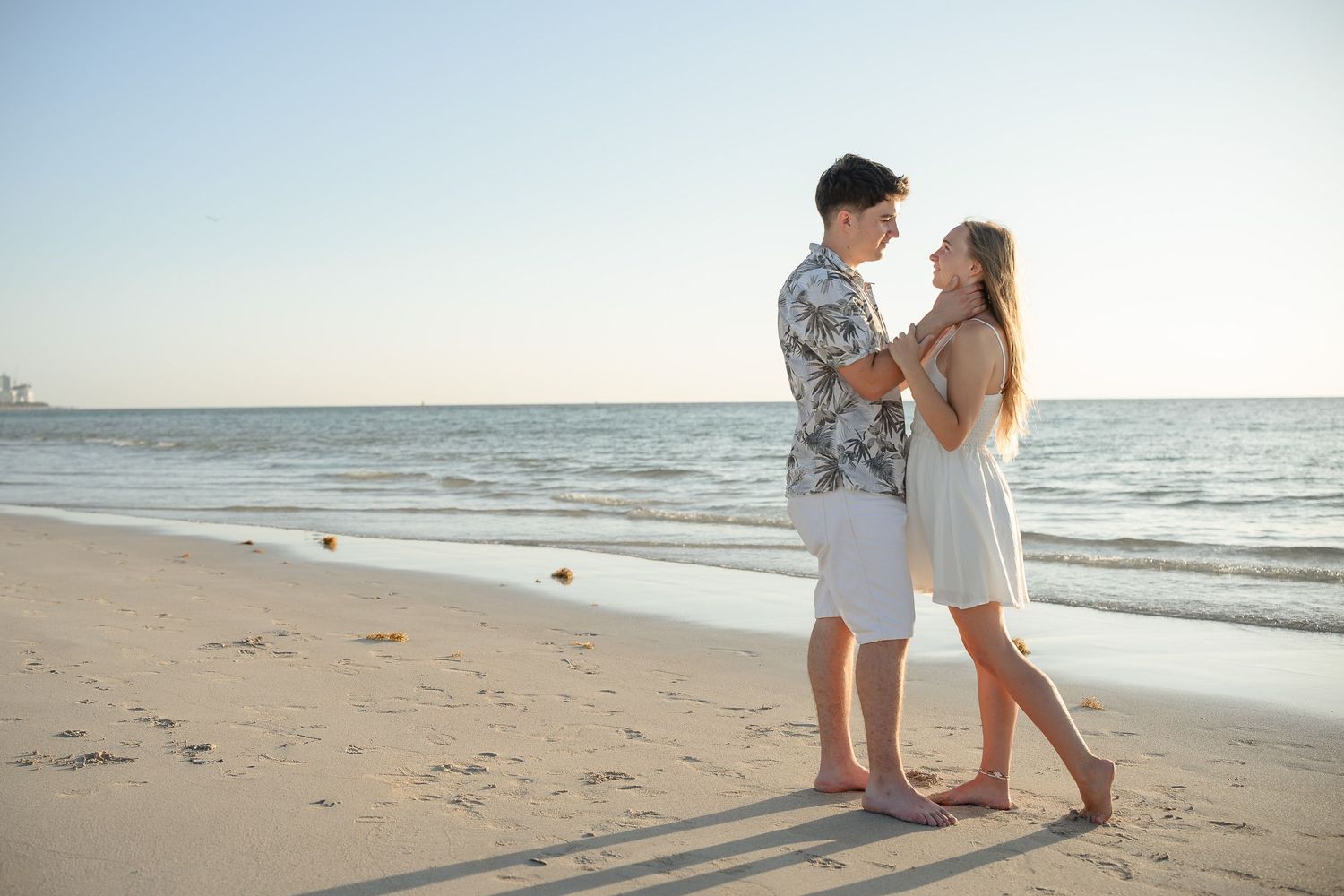 A young couple holds hands and gazes at each other romantically on a sandy beach at sunset.