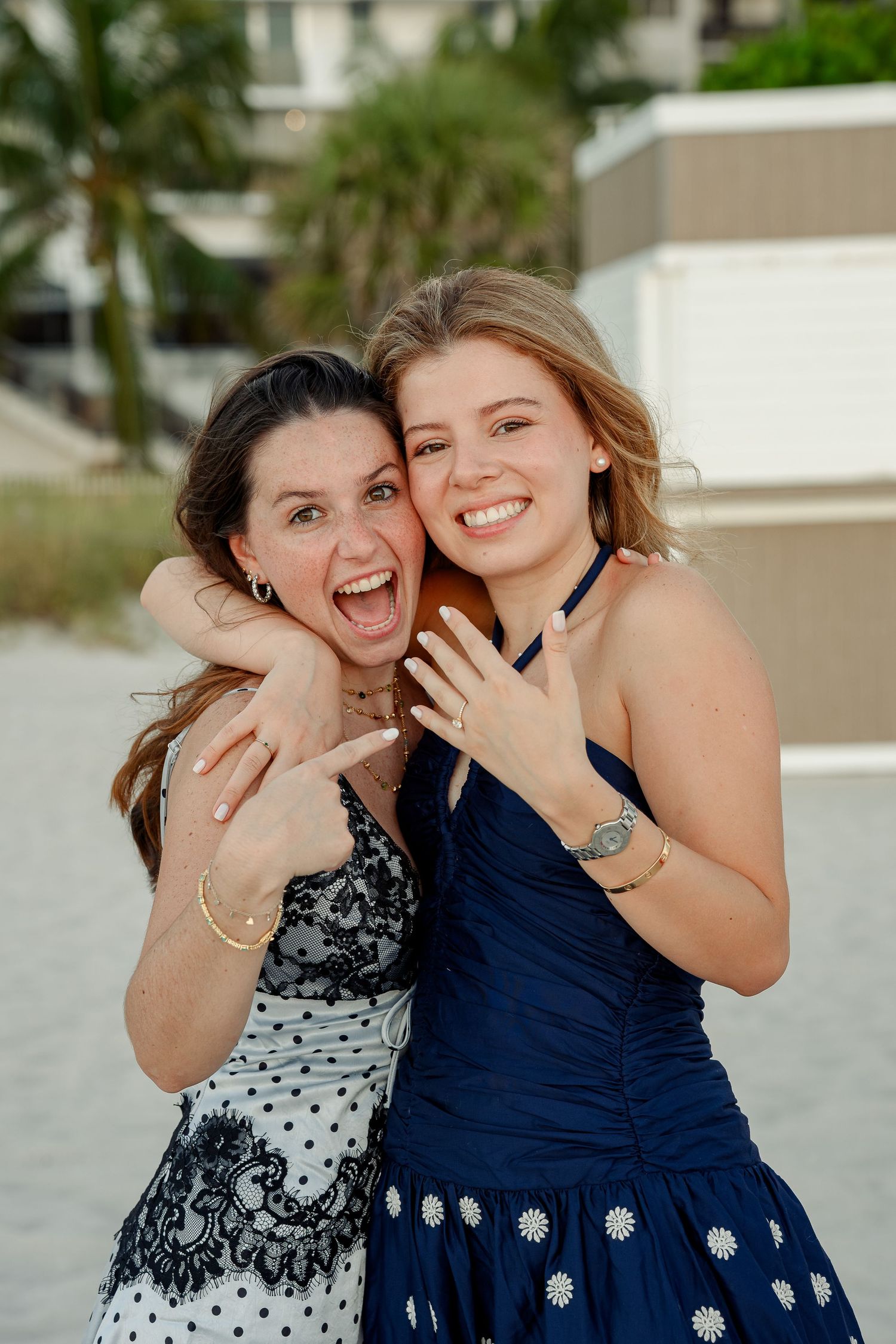 Two women in elegant dresses smiling and hugging on a beach, one showing off a ring.
