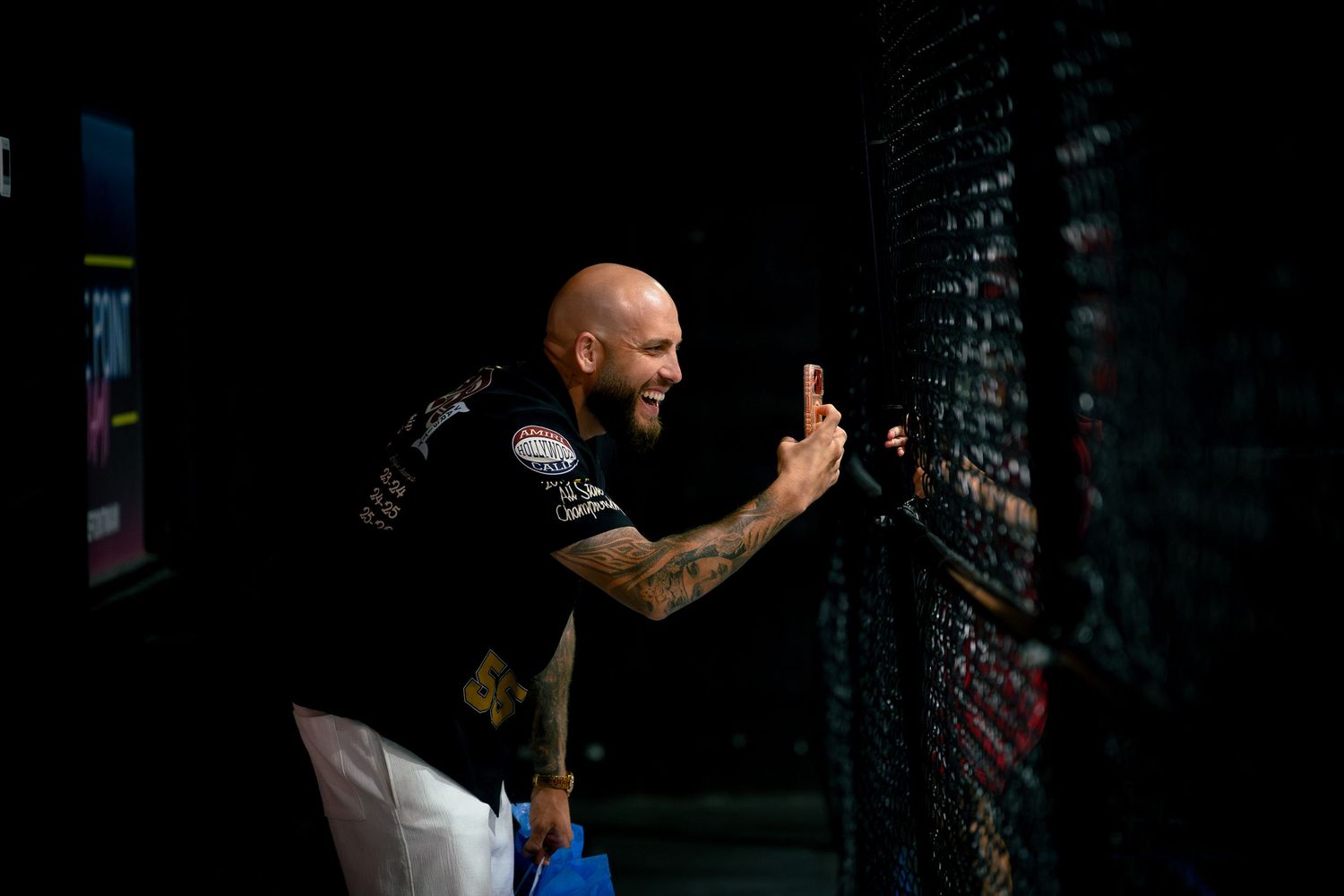 Bald tattooed MMA fighter smiling while touching octagon cage fence at UFC event in dark arena.