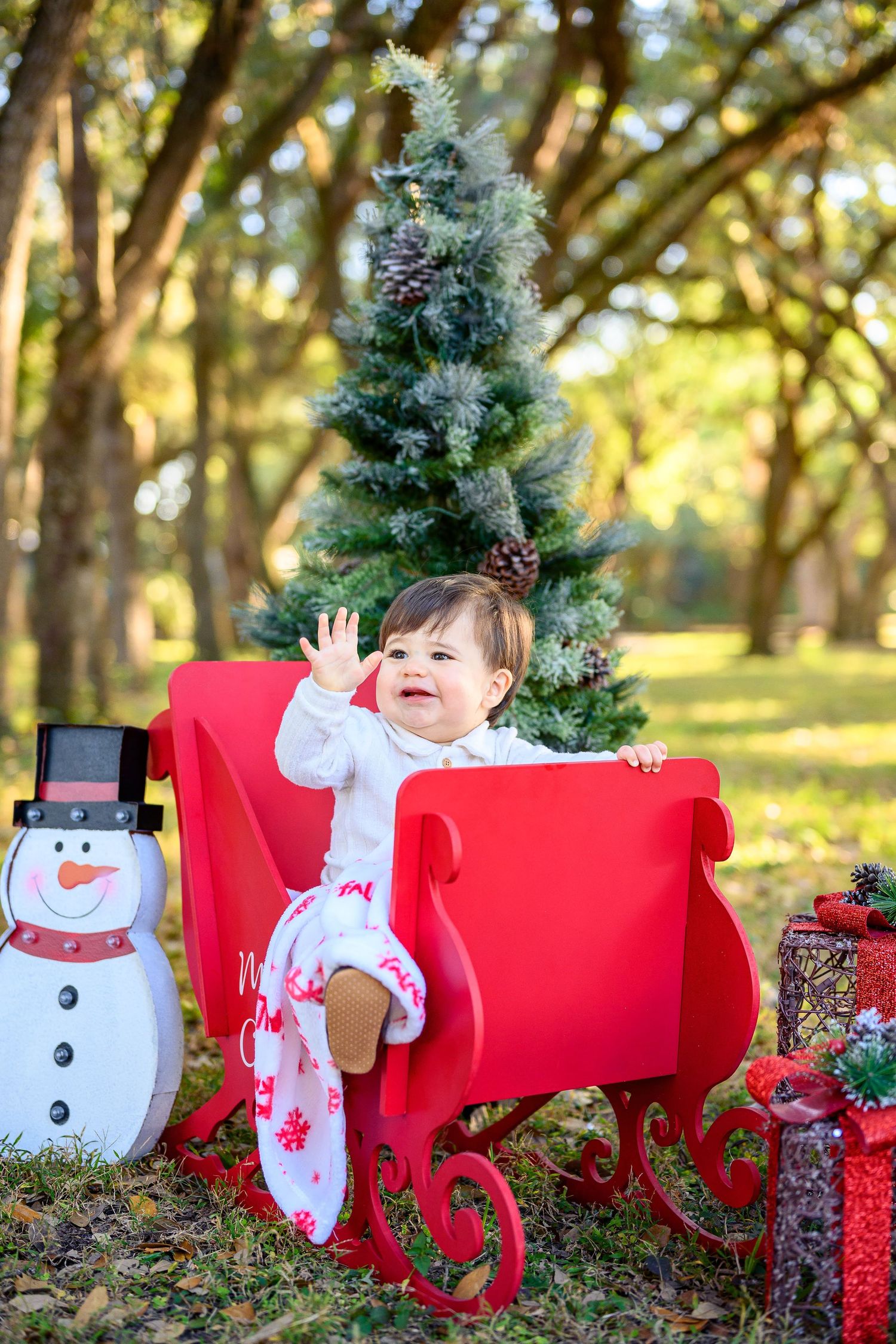 Happy baby in red sleigh waves beside snowman and Christmas tree in sunny park.