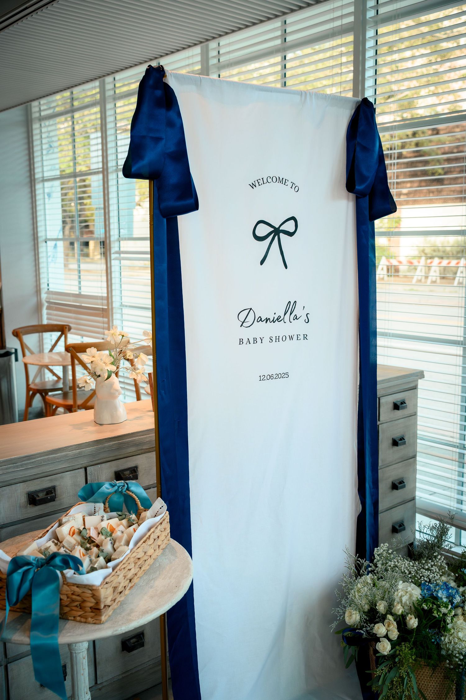 White baby shower backdrop with navy blue bow logo and accents displayed near window with floral and basket decorations.