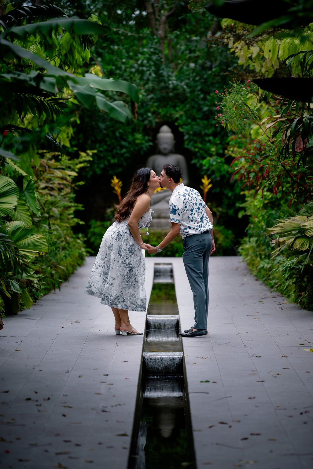 Couple sharing a romantic kiss in a lush tropical garden pathway with a Buddha statue backdrop.