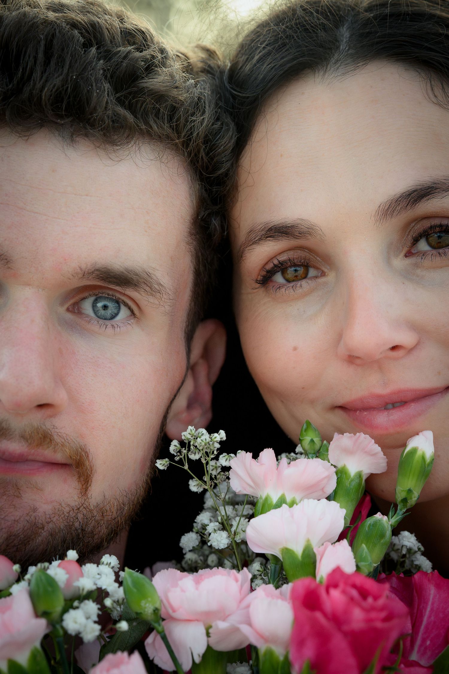 Close-up portrait of a couple with blue and brown eyes holding pink carnations and baby's breath flowers.