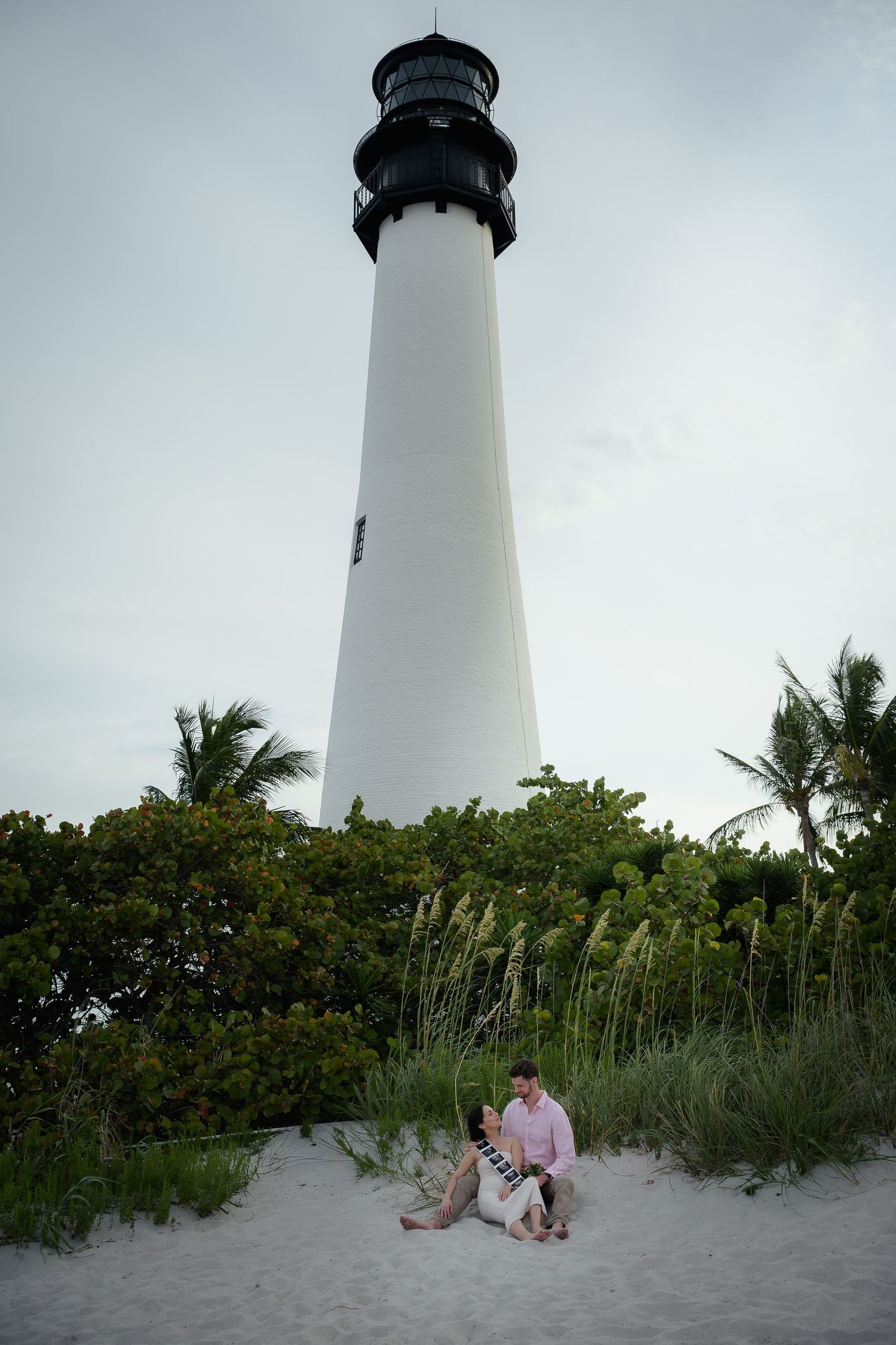 Couple sitting on sandy beach dunes in front of tall white Cape Florida Lighthouse surrounded by tropical palms.