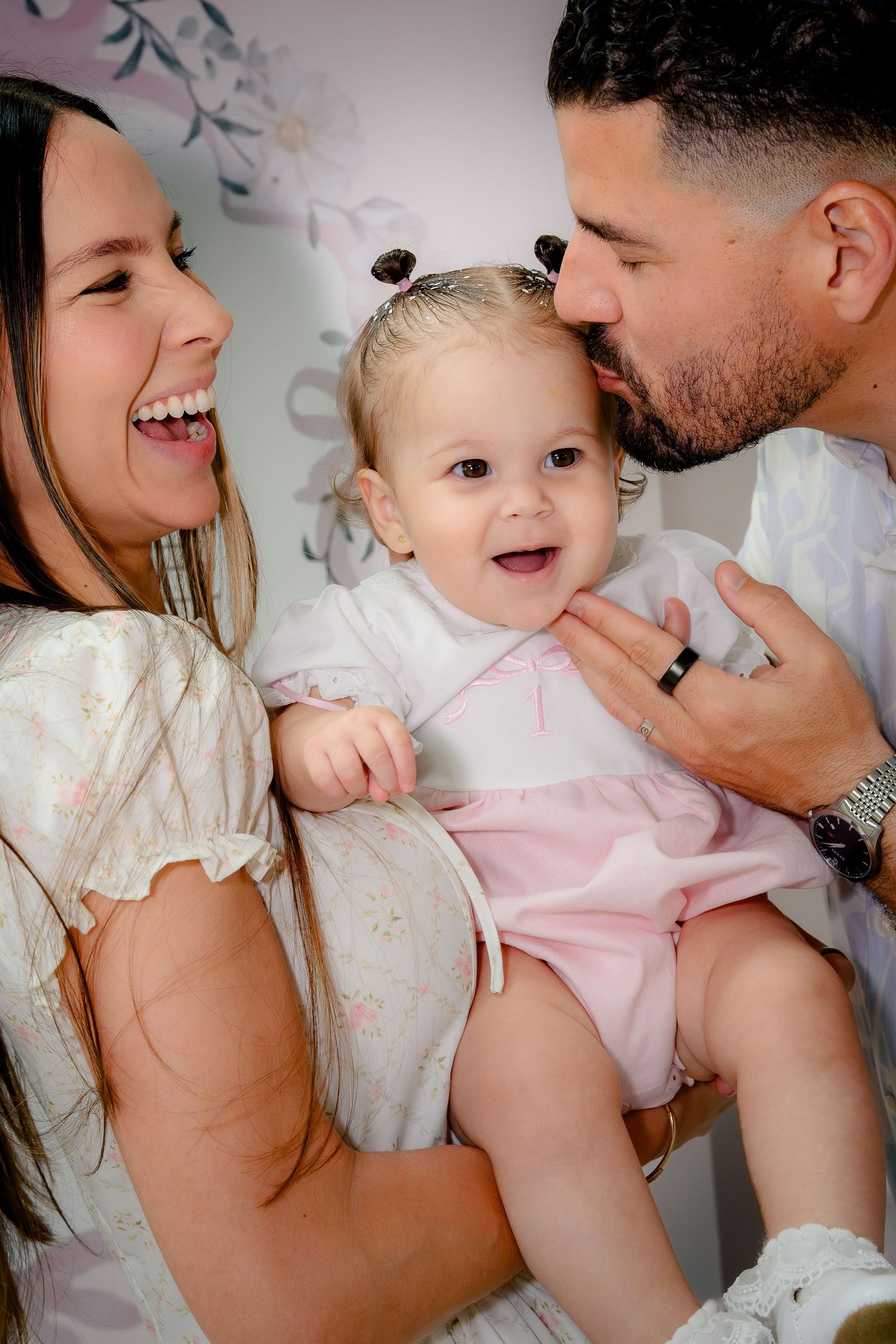 Happy family portrait with parents kissing and holding their smiling baby girl in a pink outfit.