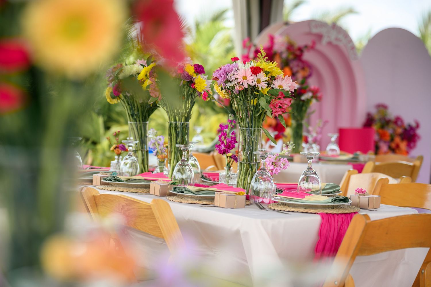Colorful floral centerpieces with pink and orange flowers adorn elegant table settings at an outdoor event.