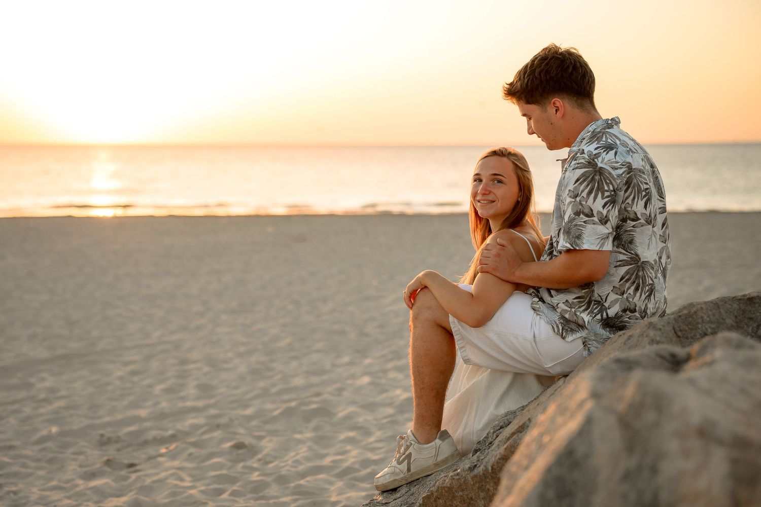 Couple sitting on rocks at sunset beach, smiling romantically in golden light.