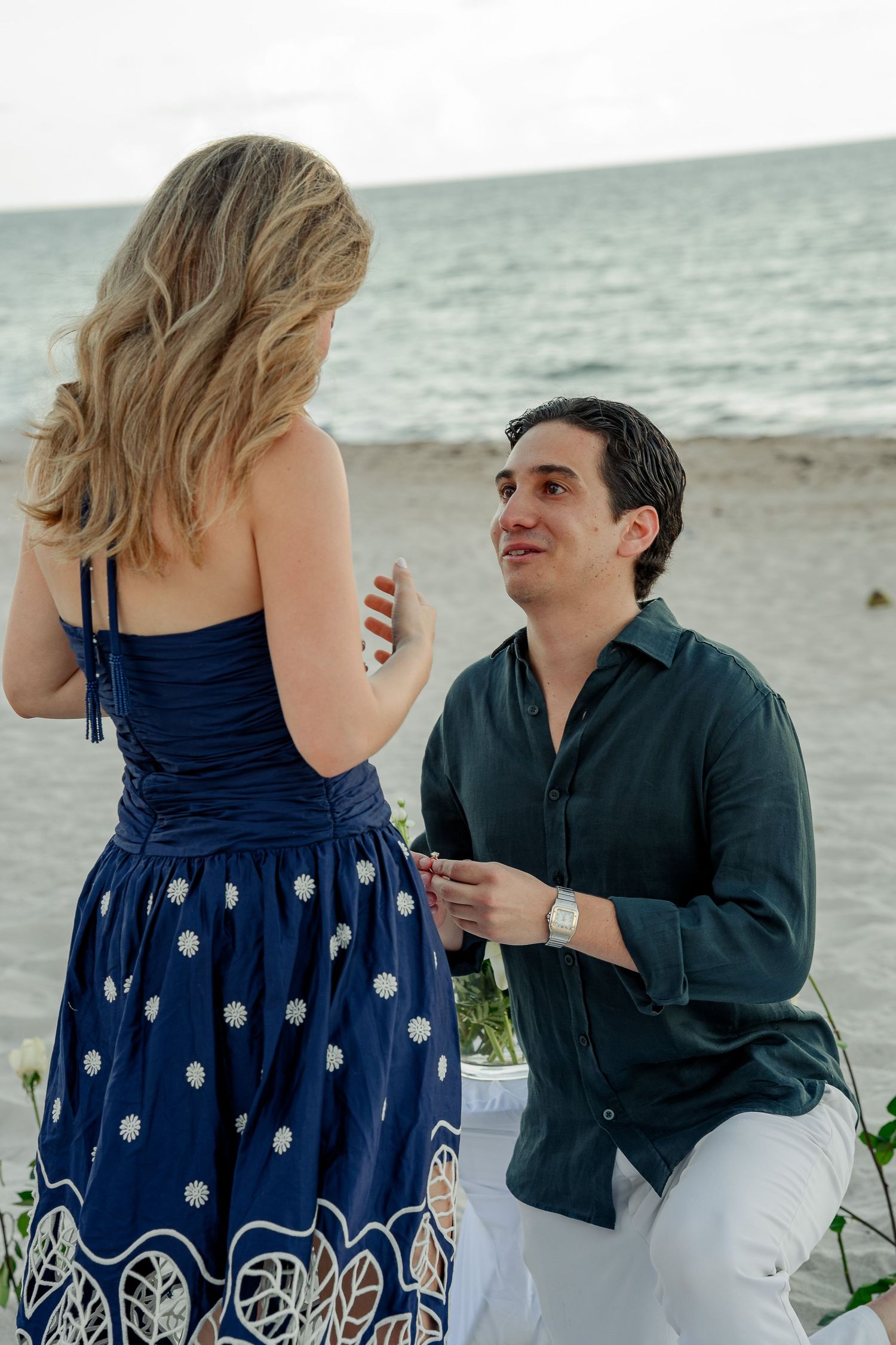 Man kneeling on beach proposing to woman in blue floral dress with ocean backdrop.