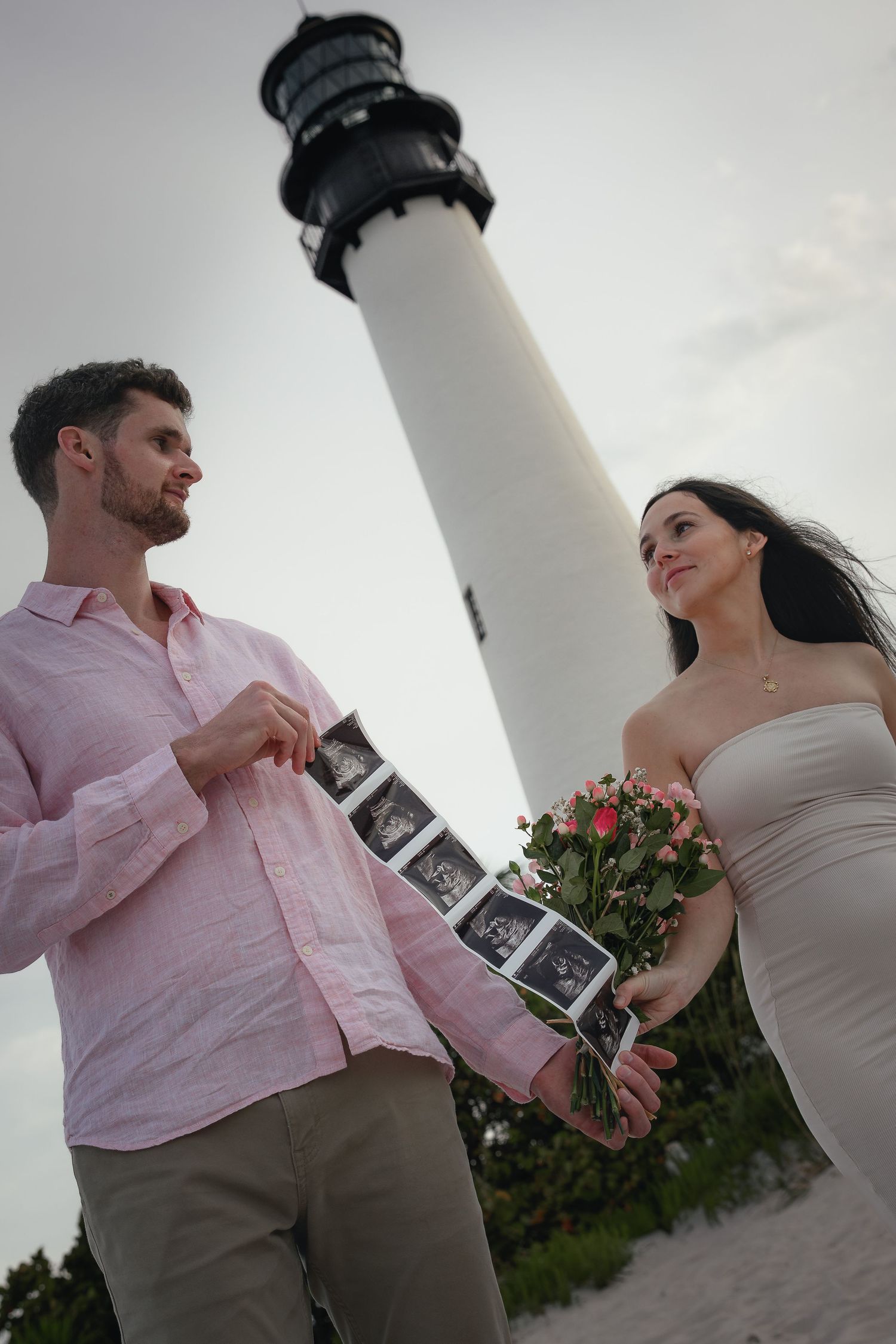 Couple holding ultrasound photos and roses by a lighthouse, announcing pregnancy.