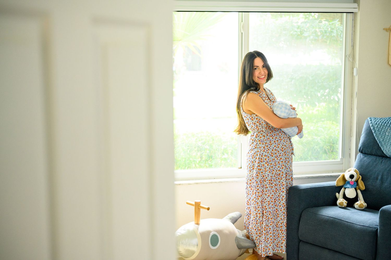 New mother standing by a sunlit window holding her newborn baby in a floral maxi dress.
