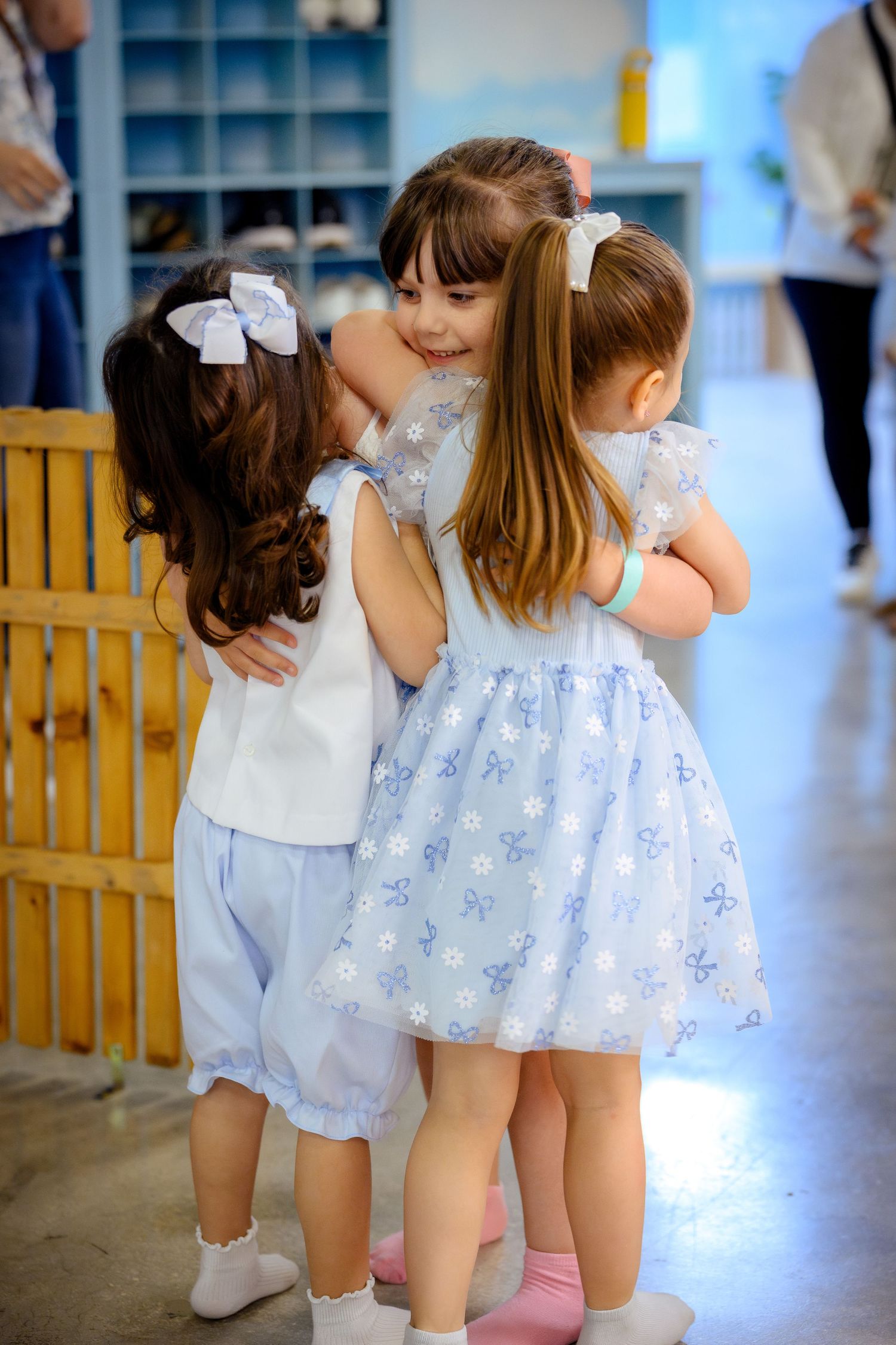Three little girls in pastel dresses sharing a group hug in a bright classroom setting.