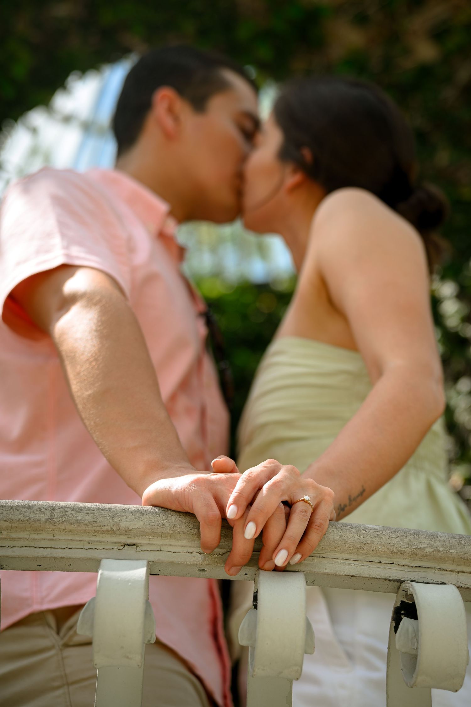 Couple kissing over a white fence railing, hands touching with an engagement ring visible in the foreground.