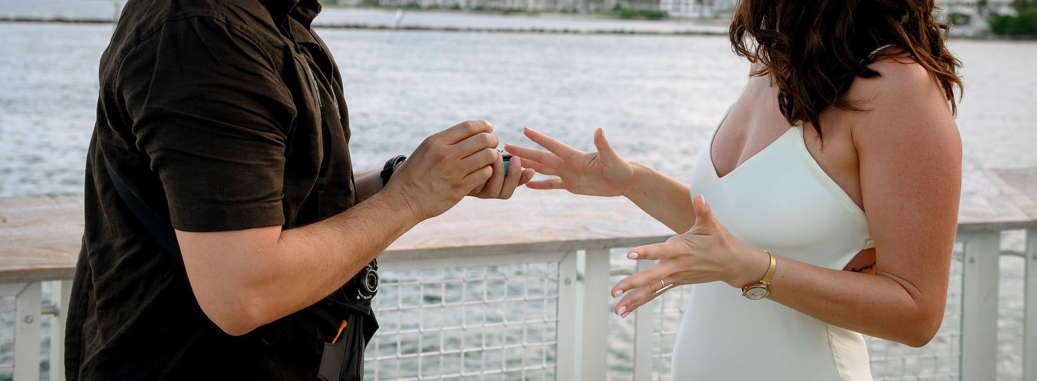 A couple exchanges rings during an outdoor waterfront proposal, with the woman in a white dress accepting.