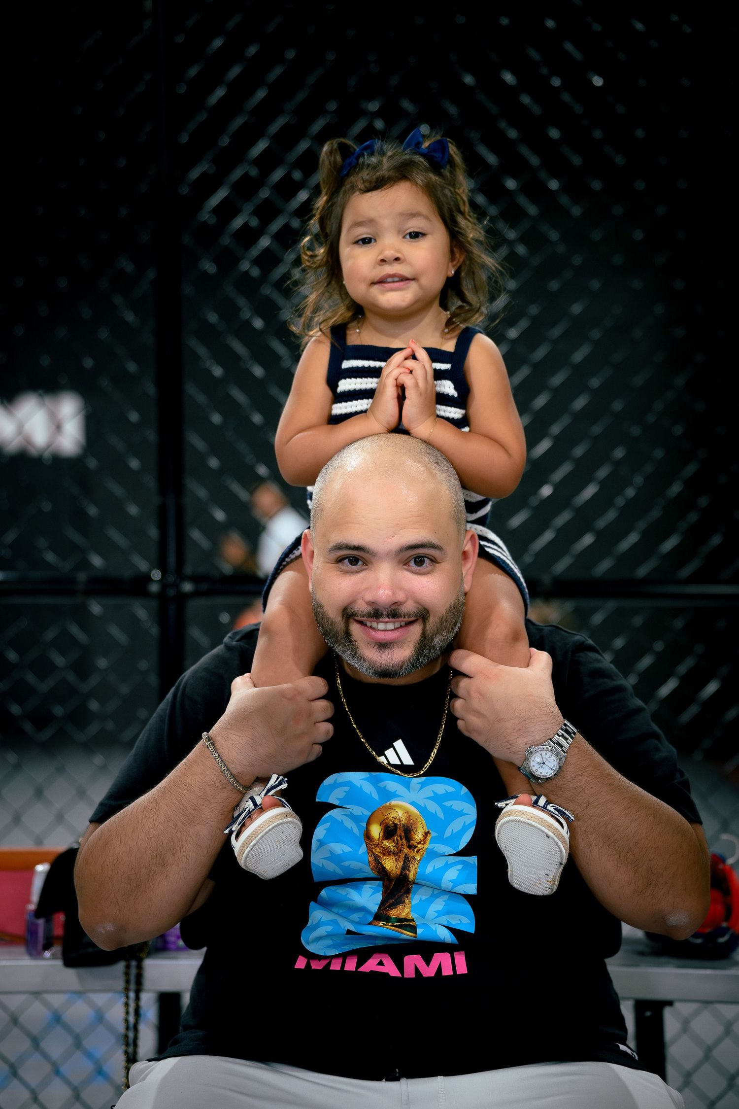 A bald man giving thumbs up wears a FIFA World Cup Miami shirt while a young girl sits on his shoulders.