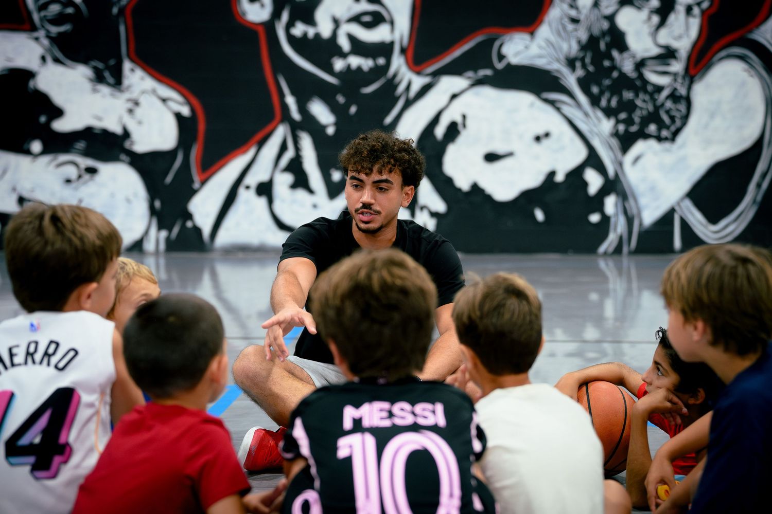 Coach talks to youth basketball campers in front of a mural, one child wearing a Messi jersey.