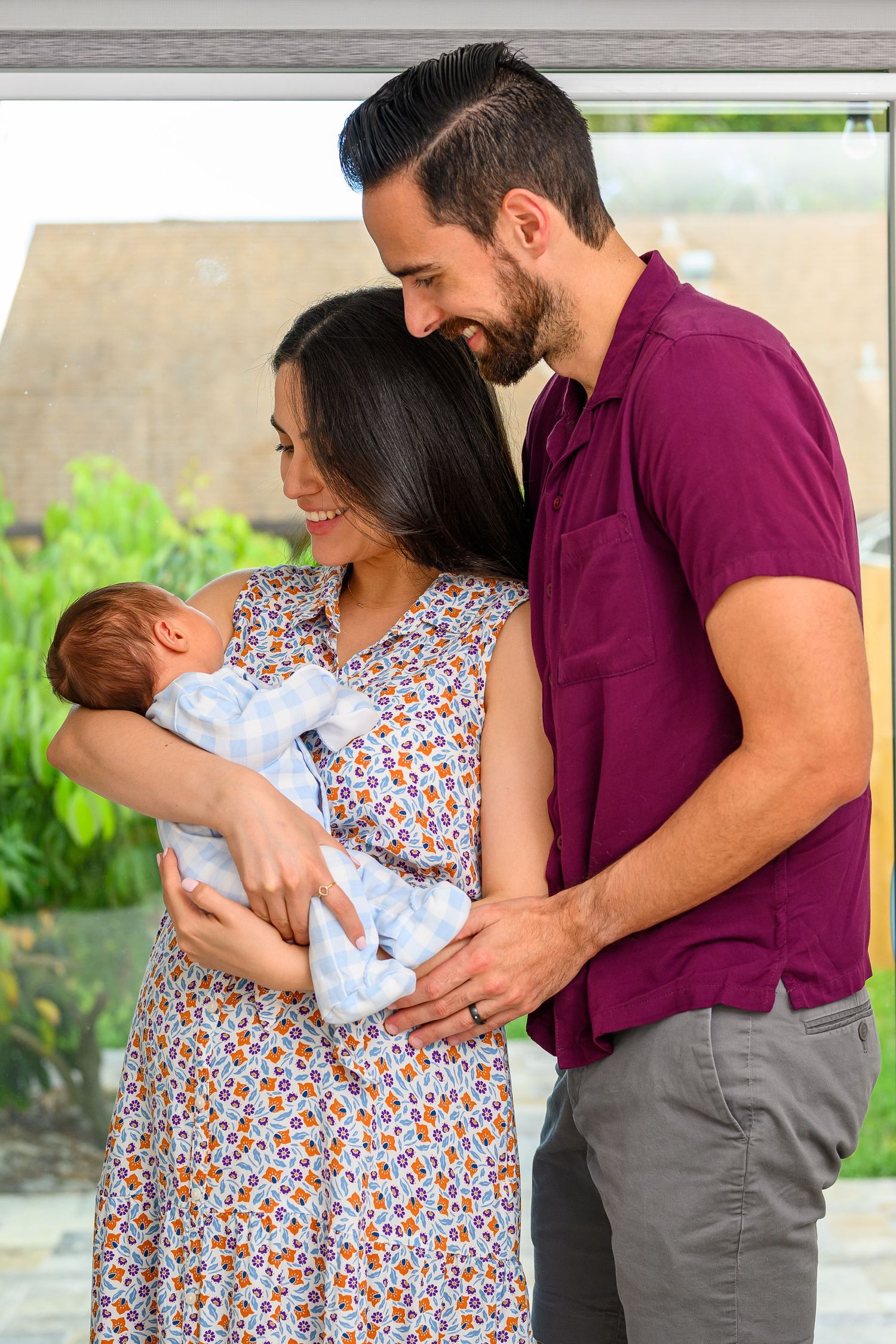 Smiling couple admiring their newborn baby together near a bright window with green garden views.