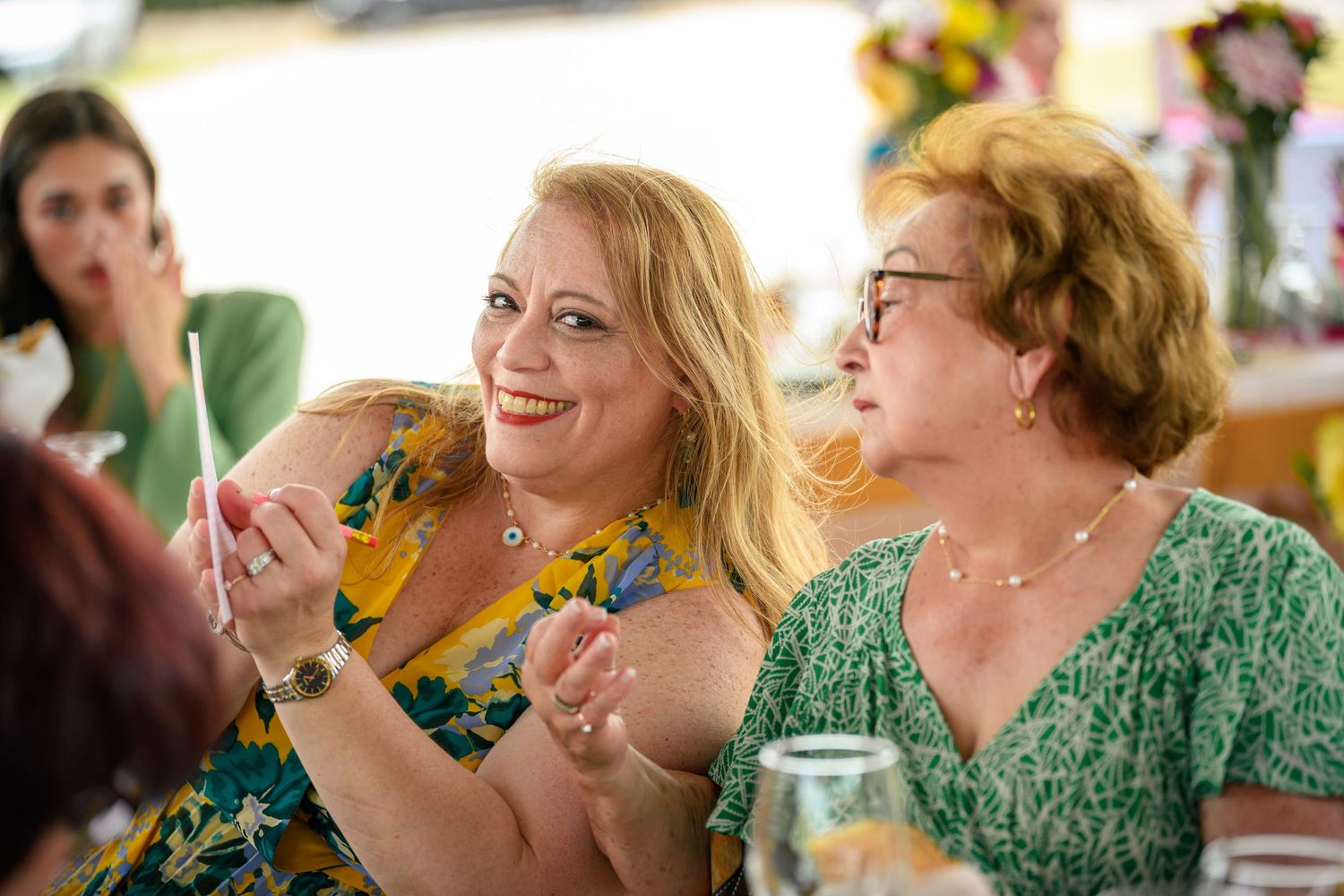 Two people in colorful floral dresses share a laugh while enjoying drinks at a social gathering.