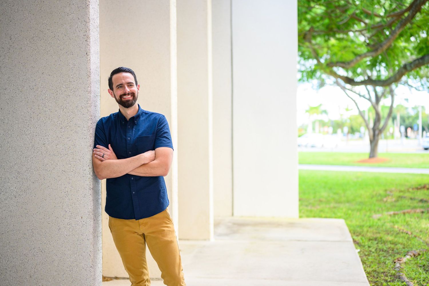 Confident smiling man with beard in navy blue shirt posing outdoors against a white building wall.