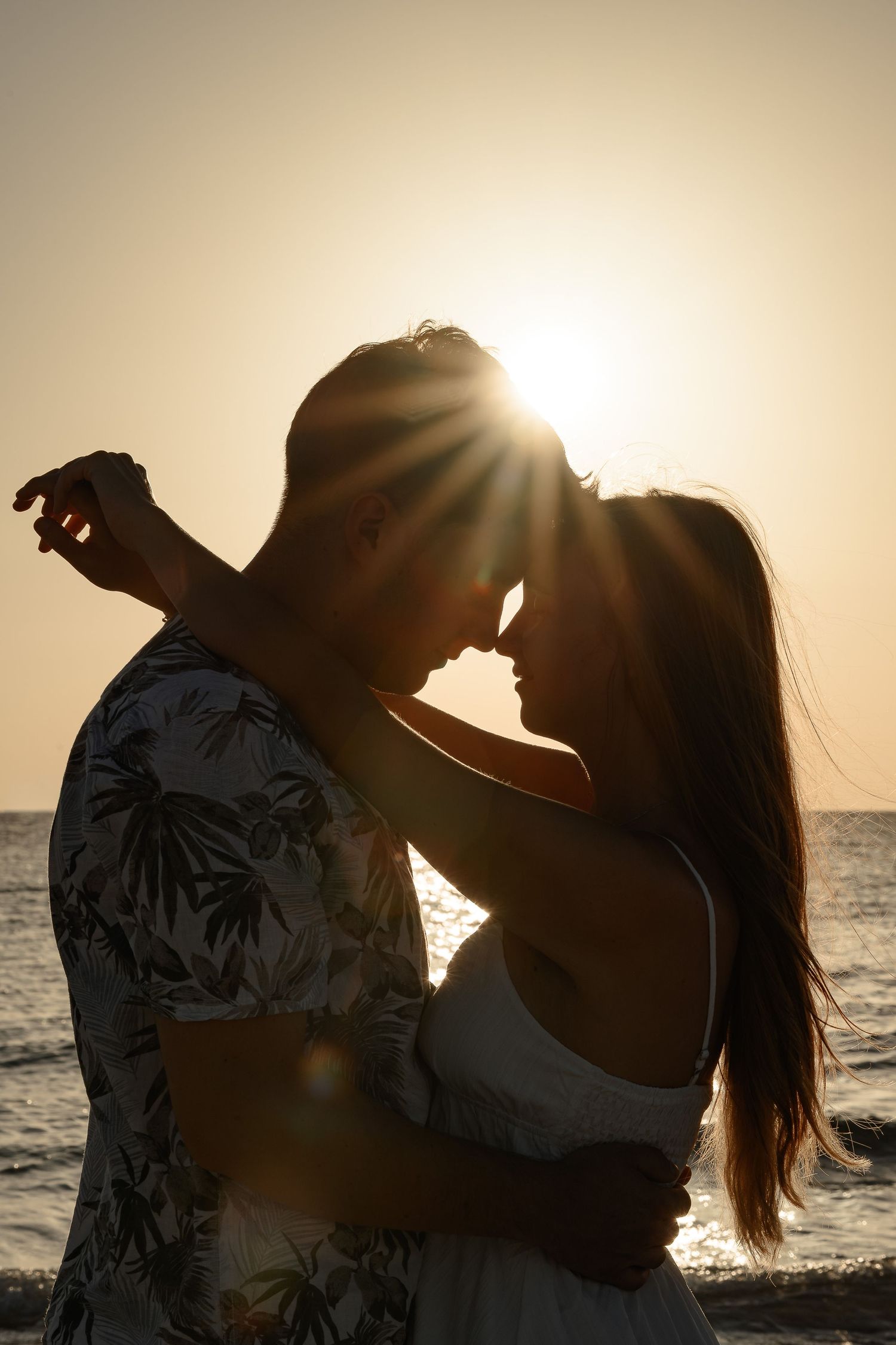 Romantic couple silhouetted against a golden sunset at the beach, leaning in for a kiss by the ocean.