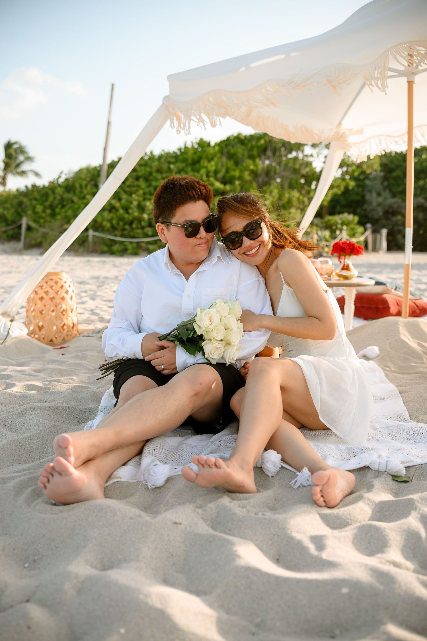 Couple sitting on beach blanket under white umbrella, holding white roses, wearing sunglasses, enjoying a romantic beach picnic.