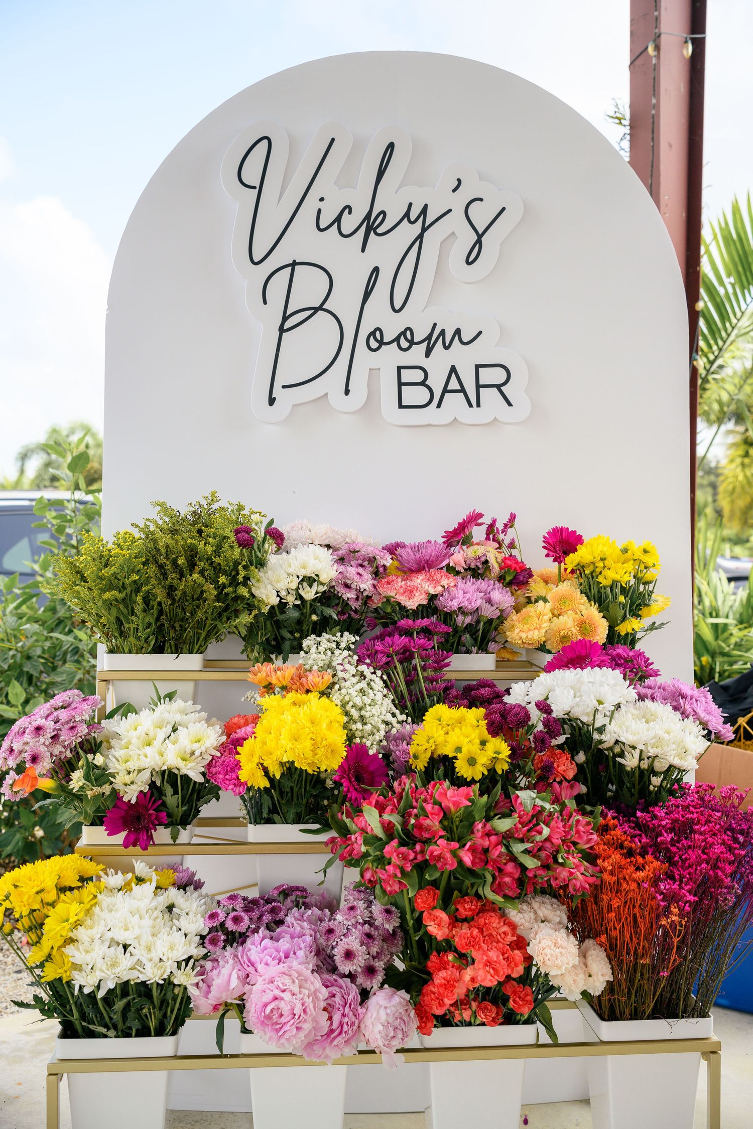 A colorful flower stand with 'Vicky's Bloom Bar' sign displaying vibrant pink, yellow, and white fresh flowers.