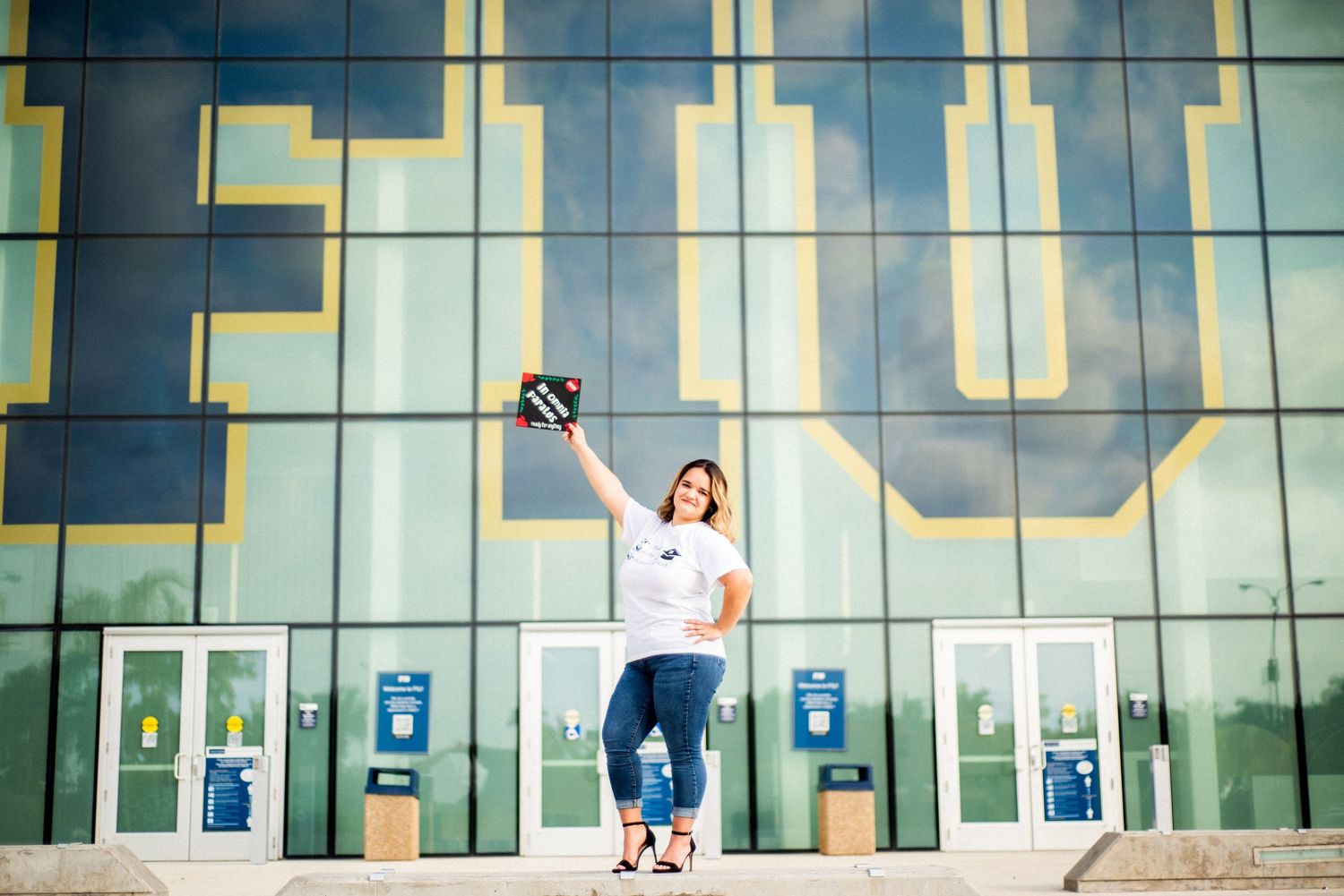 Joyful woman in white shirt and jeans raising graduation cap outside a large glass university building.