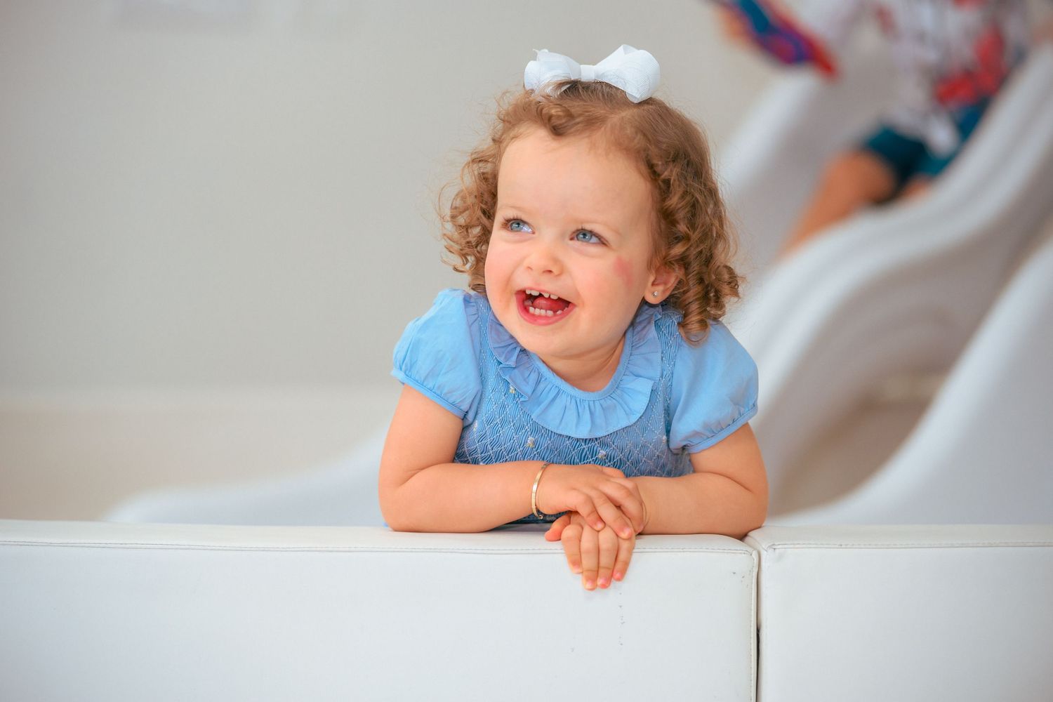 Smiling toddler in blue dress leaning on white surface during playful photoshoot.