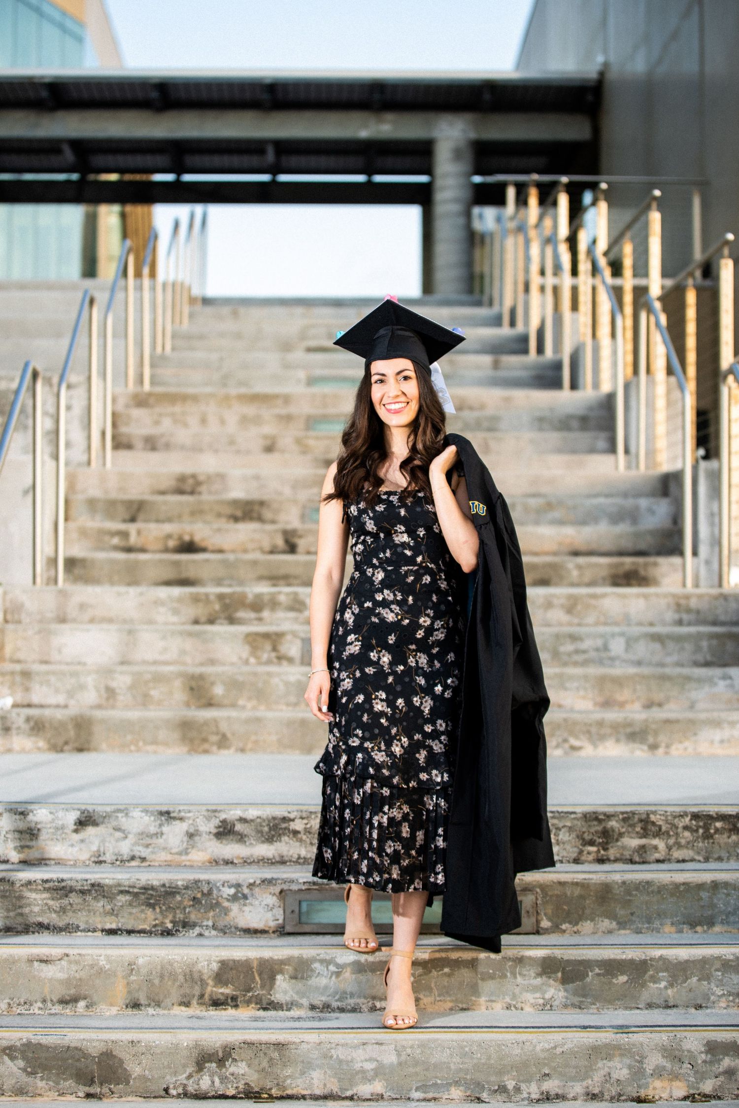 Smiling graduate in floral dress and black cap holds gown on outdoor university steps.