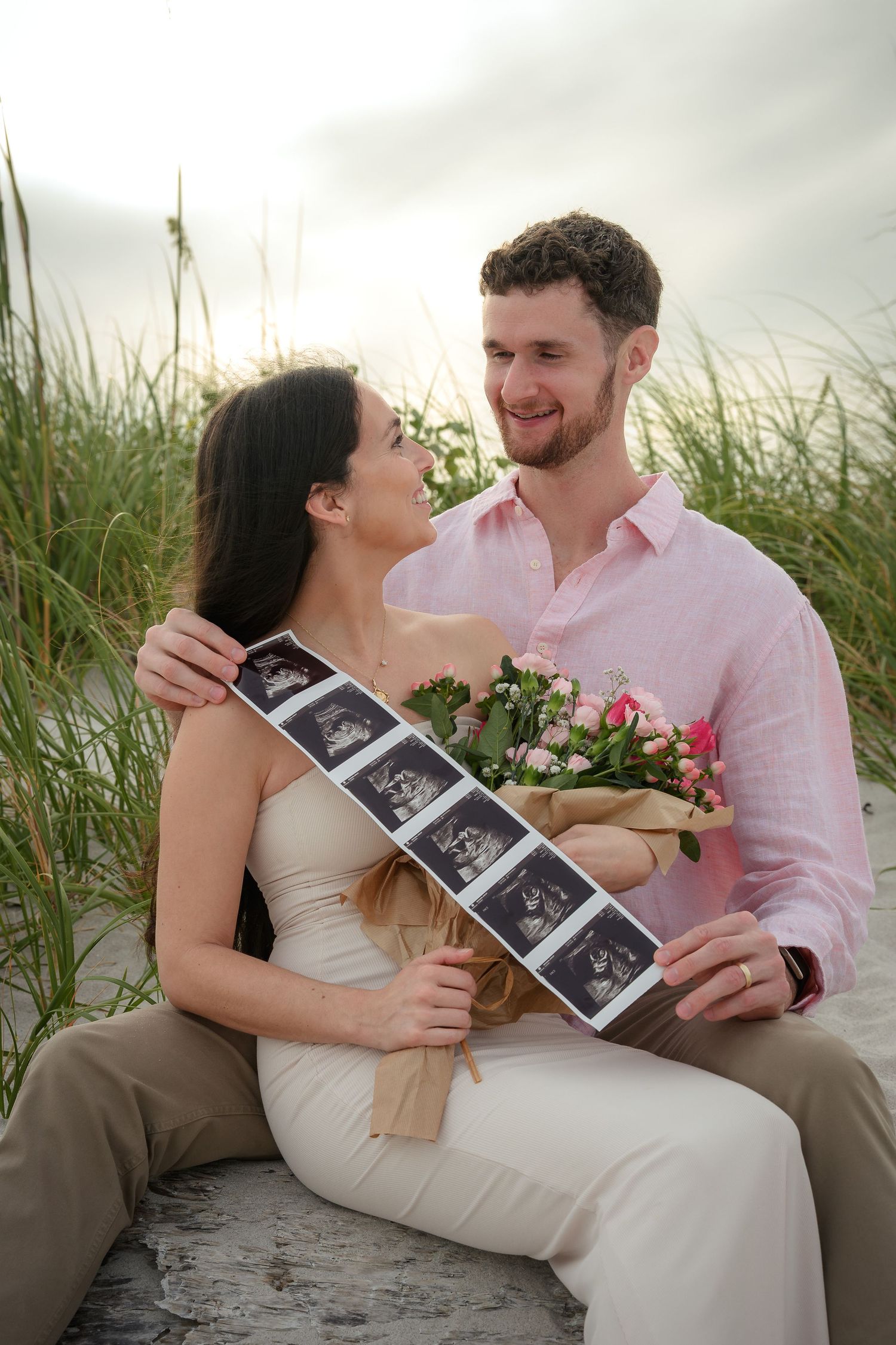 Happy couple sitting on beach sand dunes, holding ultrasound photos and pink flowers, sharing a romantic pregnancy announcement moment.