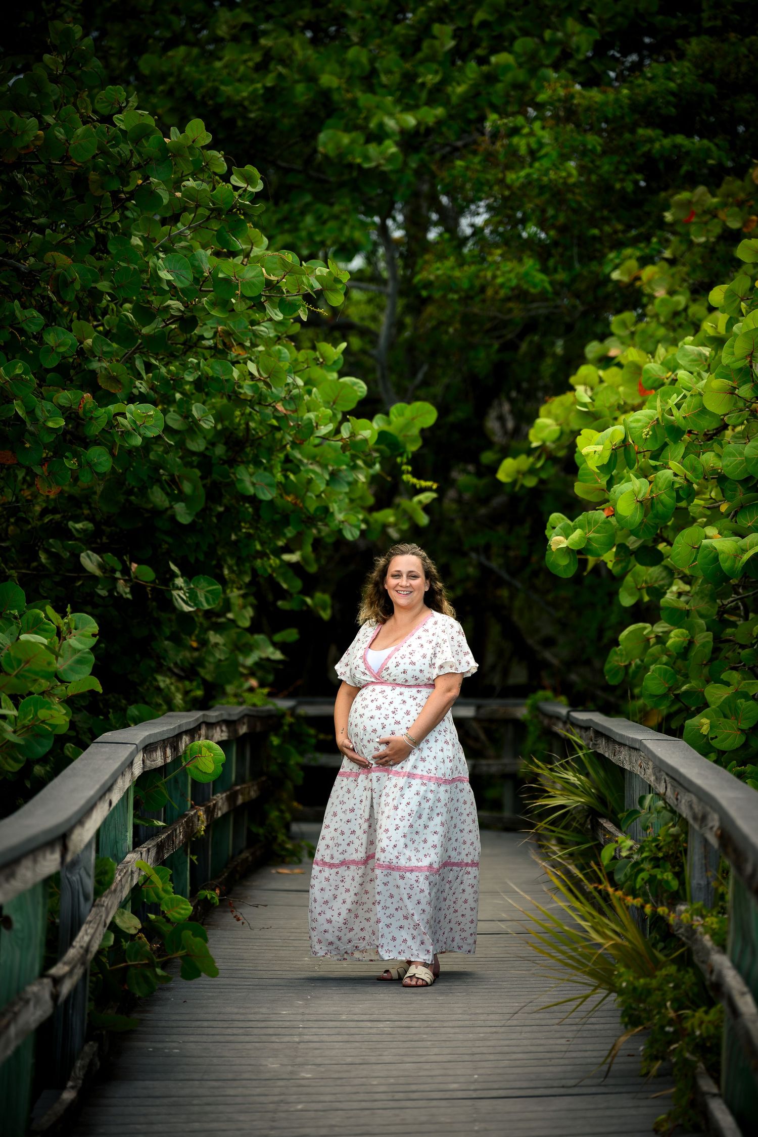 Pregnant woman in floral dress standing on wooden bridge surrounded by lush green tropical foliage.