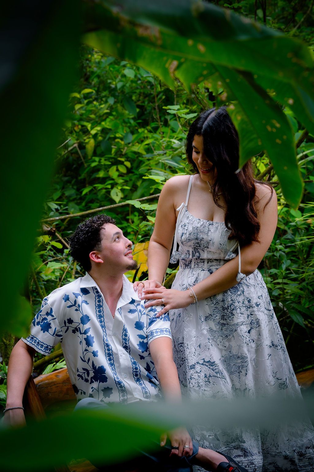 A couple shares a romantic moment in a lush tropical garden, man seated looking up at woman in floral dress.