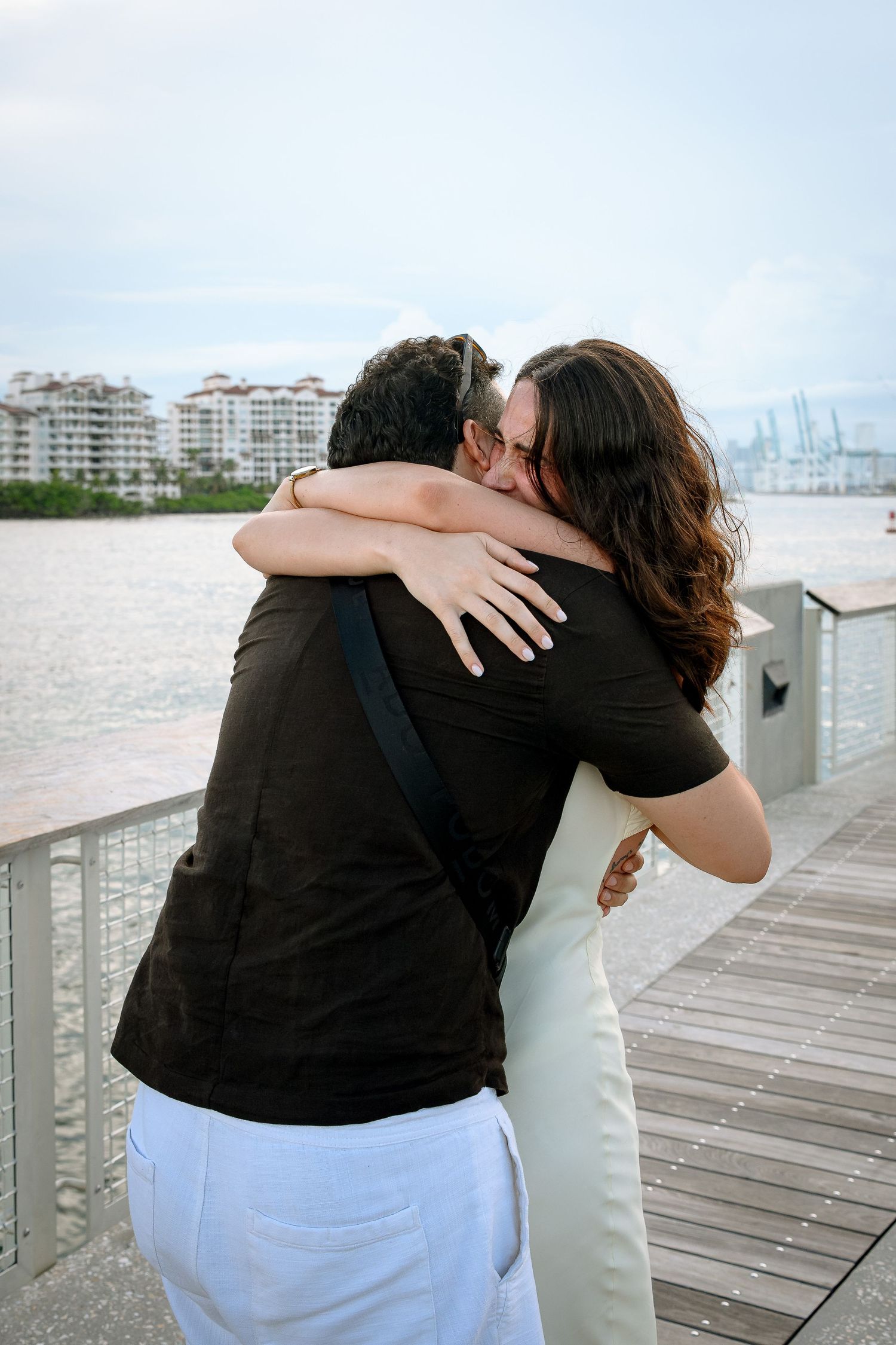 Couple embracing in a warm hug on a waterfront boardwalk with city buildings and water in the background.