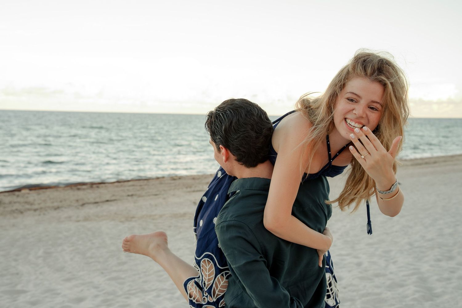 Couple laughing and embracing on a sandy beach at sunset, woman showing engagement ring.