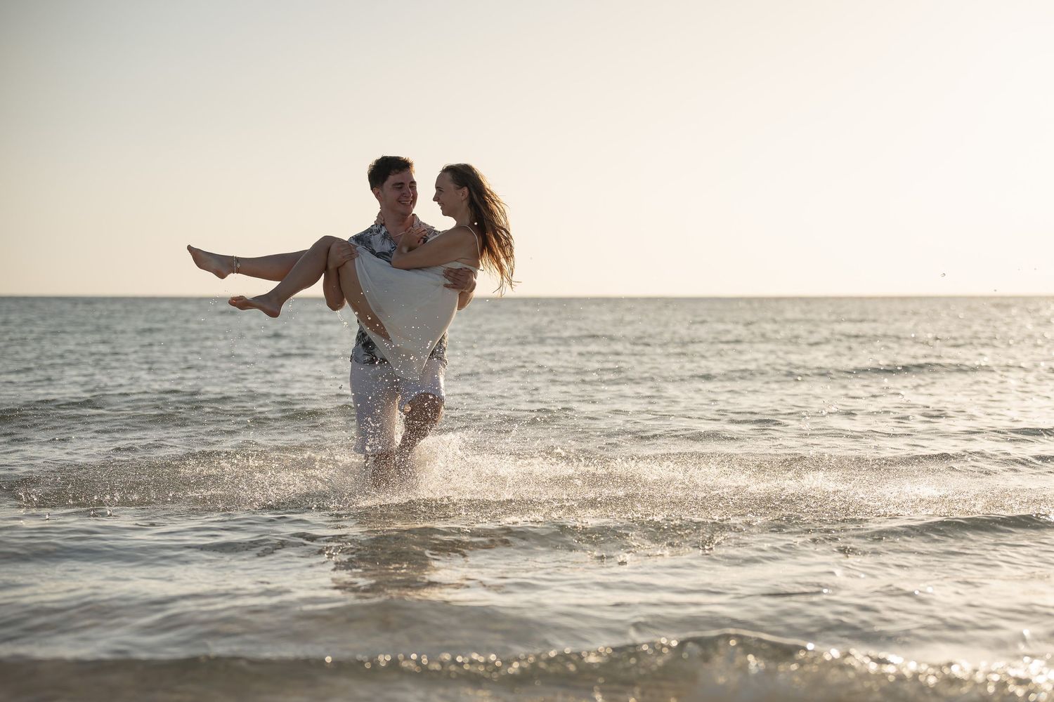 Romantic couple playing in ocean waves at sunset, man carrying woman in his arms on a beautiful sandy beach.