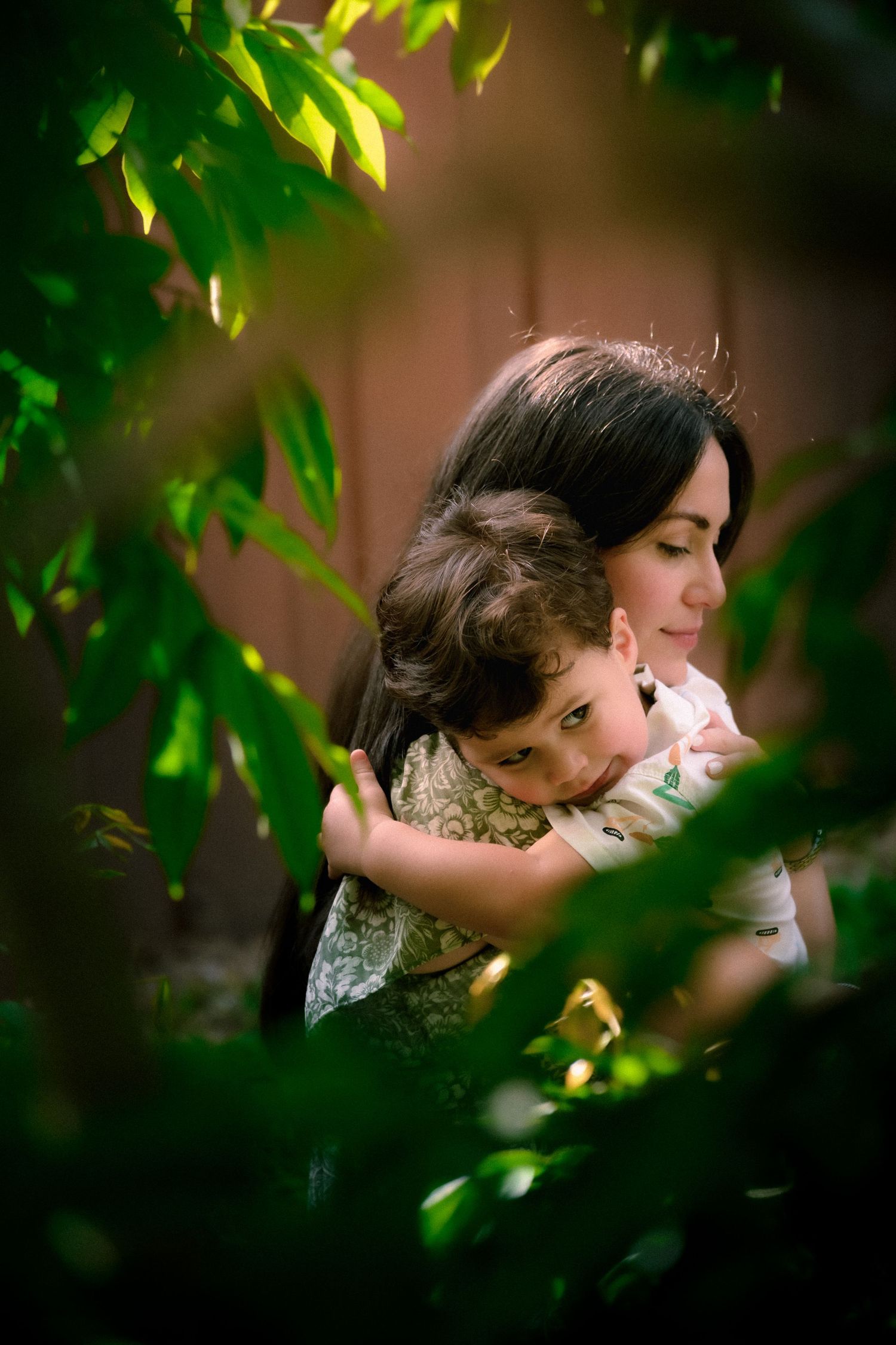 A mother tenderly holds her young child among lush green leaves in a garden setting.