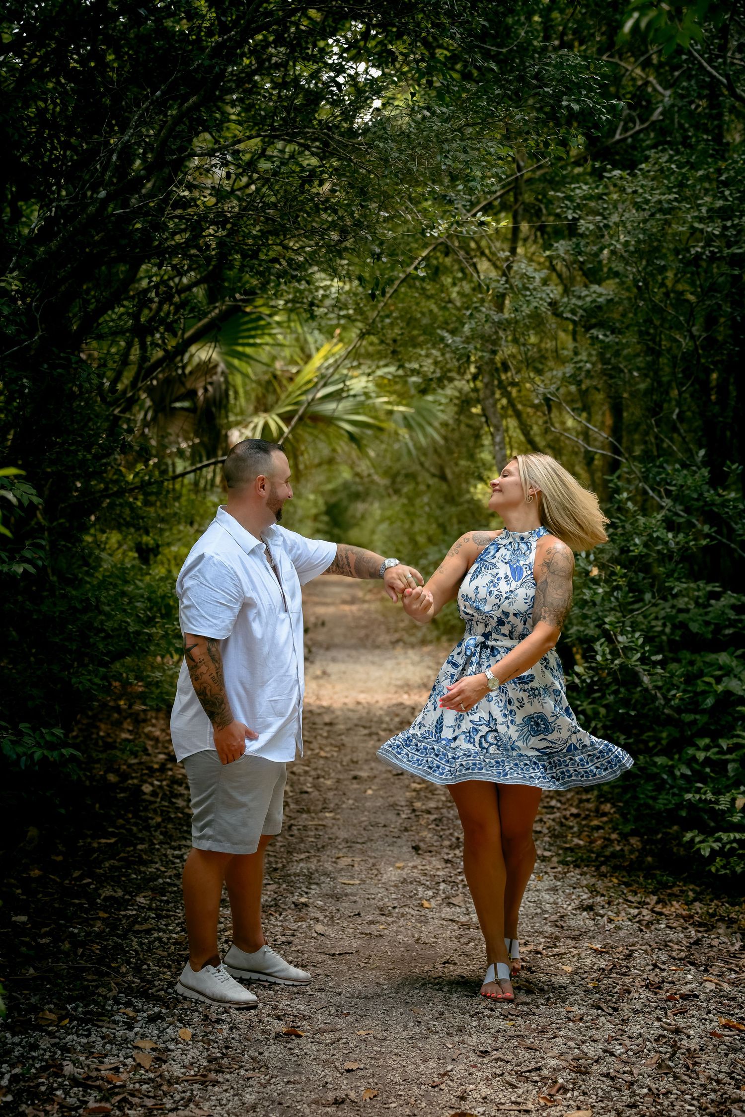 Couple holding hands and dancing on a forest path, woman in blue floral dress, man in white shirt and shorts.