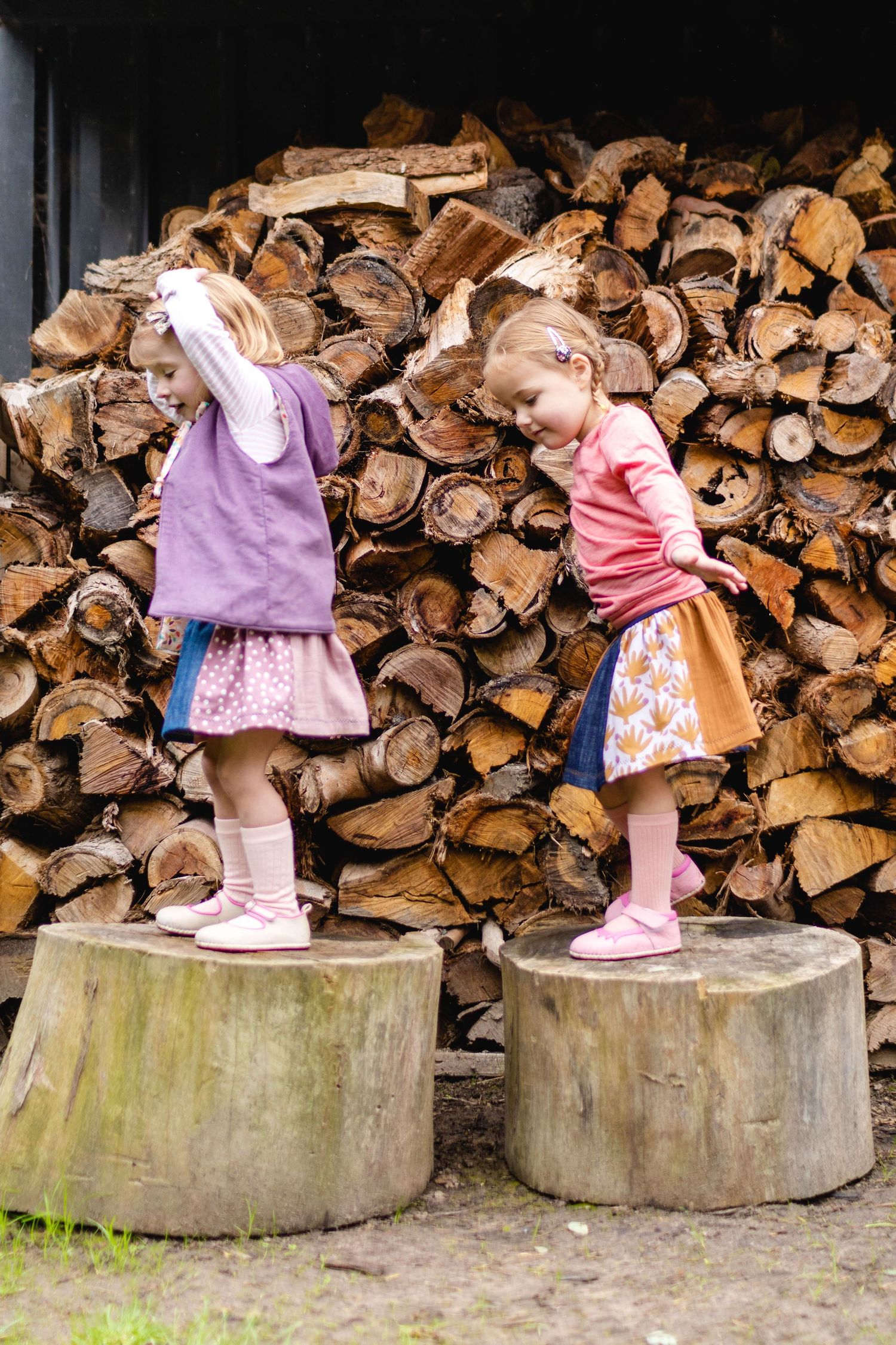 Two children playing on logs in front of stacked wood, natural outdoor lifestyle photography